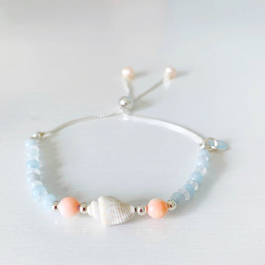the seersucker shellebration bracelet by mermaids and madeleines is a sterling silver and beaded adjustable bracelet with a shell and coral beads at the center complimented by alternating aquamarine and moonstone. this bracelet is photographed on a white surface