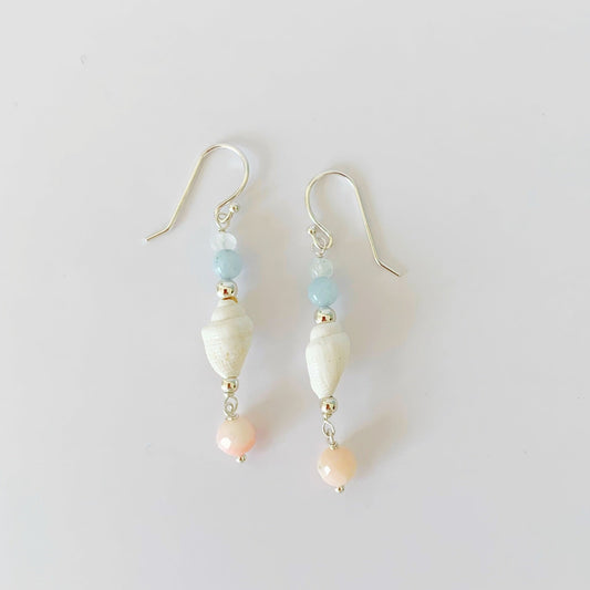 the seersucker shellebration earrings by mermaids and madeleines are a linear drop style earrings with a shell at the center complimented by moonstone, aquamarine, and with a coral bead drop at the bottom. this pair is photographed on a white surface