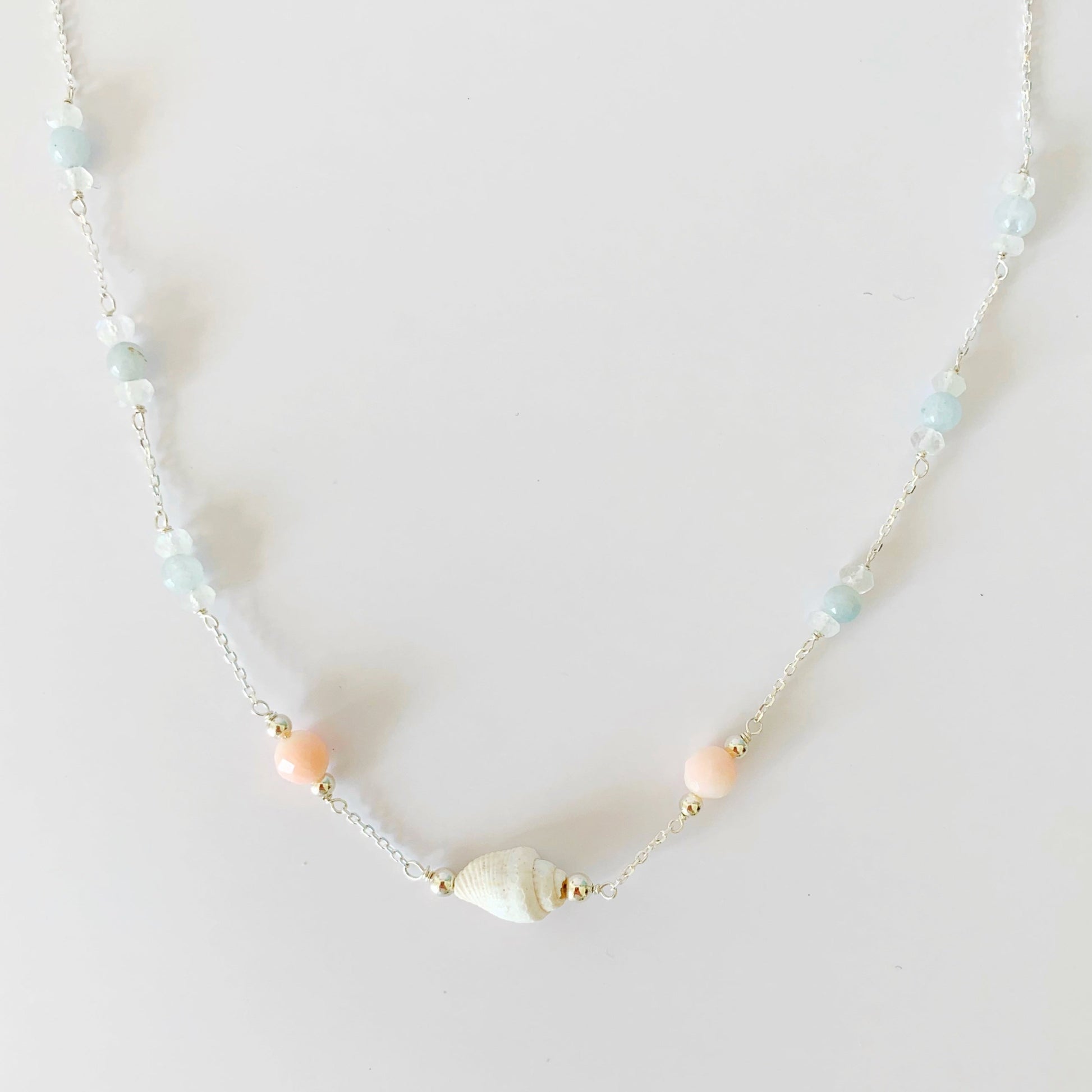 the seersucker necklace by mermaids and madeleines is a station style necklace with a shell and coral beads at the center with segments of moonstone and aquamarine along the sterling chain to the clasp. this necklace is photographed on a white surface