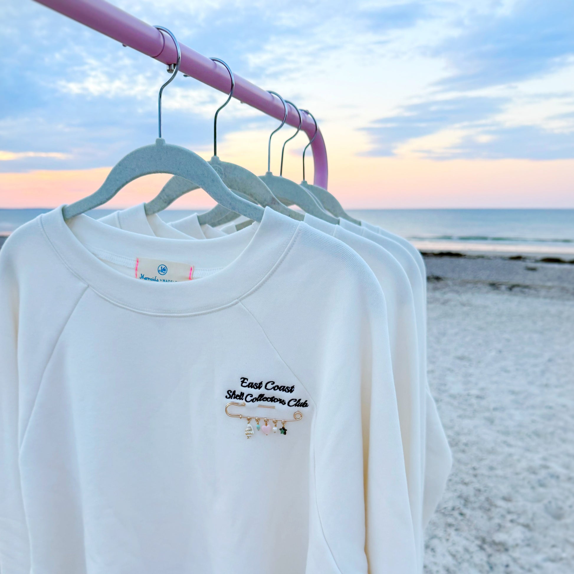 A white long-sleeve sweatshirts hanging on a light blue hanger from a pink clothing rack, set against a beach backdrop at sunset.