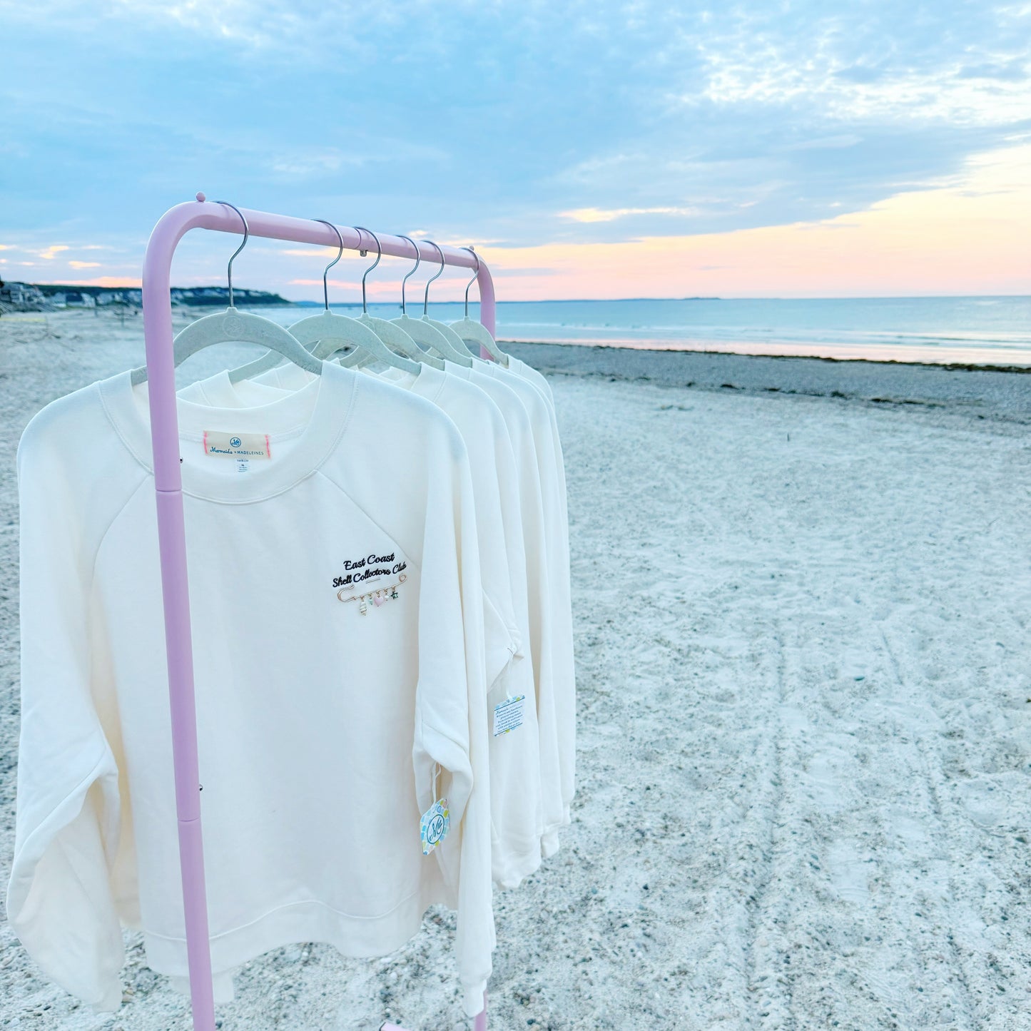 A pink rolling clothing rack on a sandy beach at sunset, holding several white long-sleeve t-shirts with small navy graphic prints on the chest.
