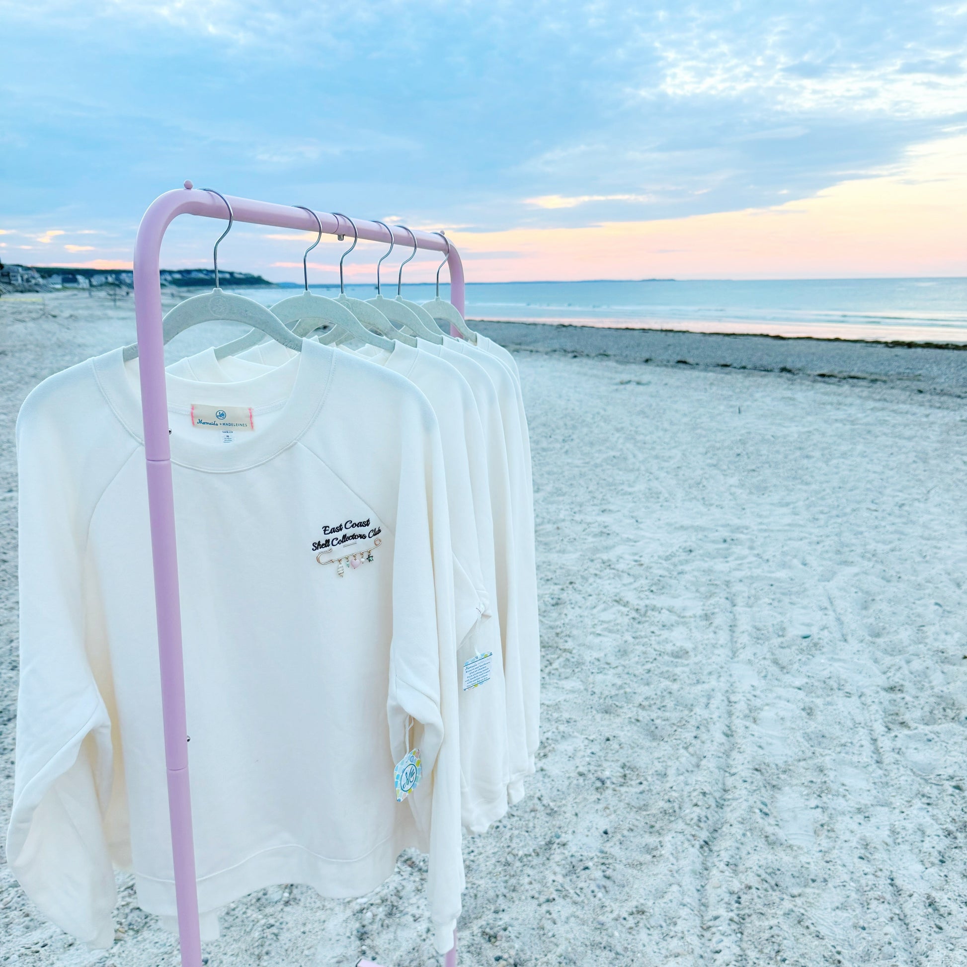 A pink rolling clothing rack on a sandy beach at sunset, holding several white long-sleeve t-shirts with small navy graphic prints on the chest.