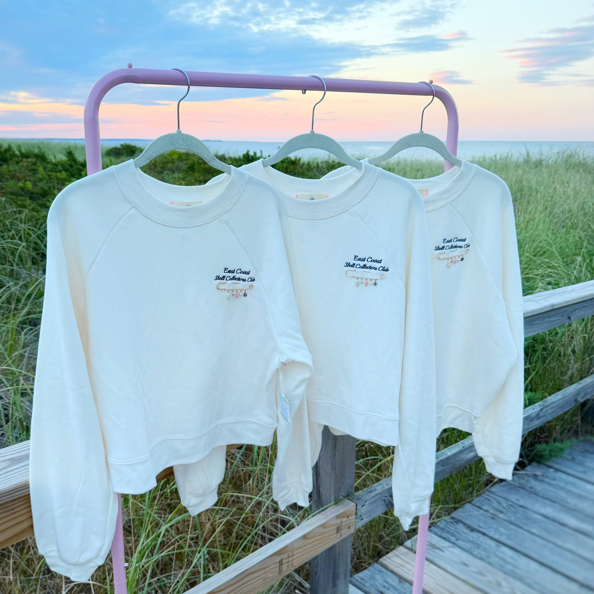 Three white crewneck sweatshirts with small navy blue embroidered text hang on a pink metal clothing rack outdoors on a wooden boardwalk with grass and a sunset sky in the background.