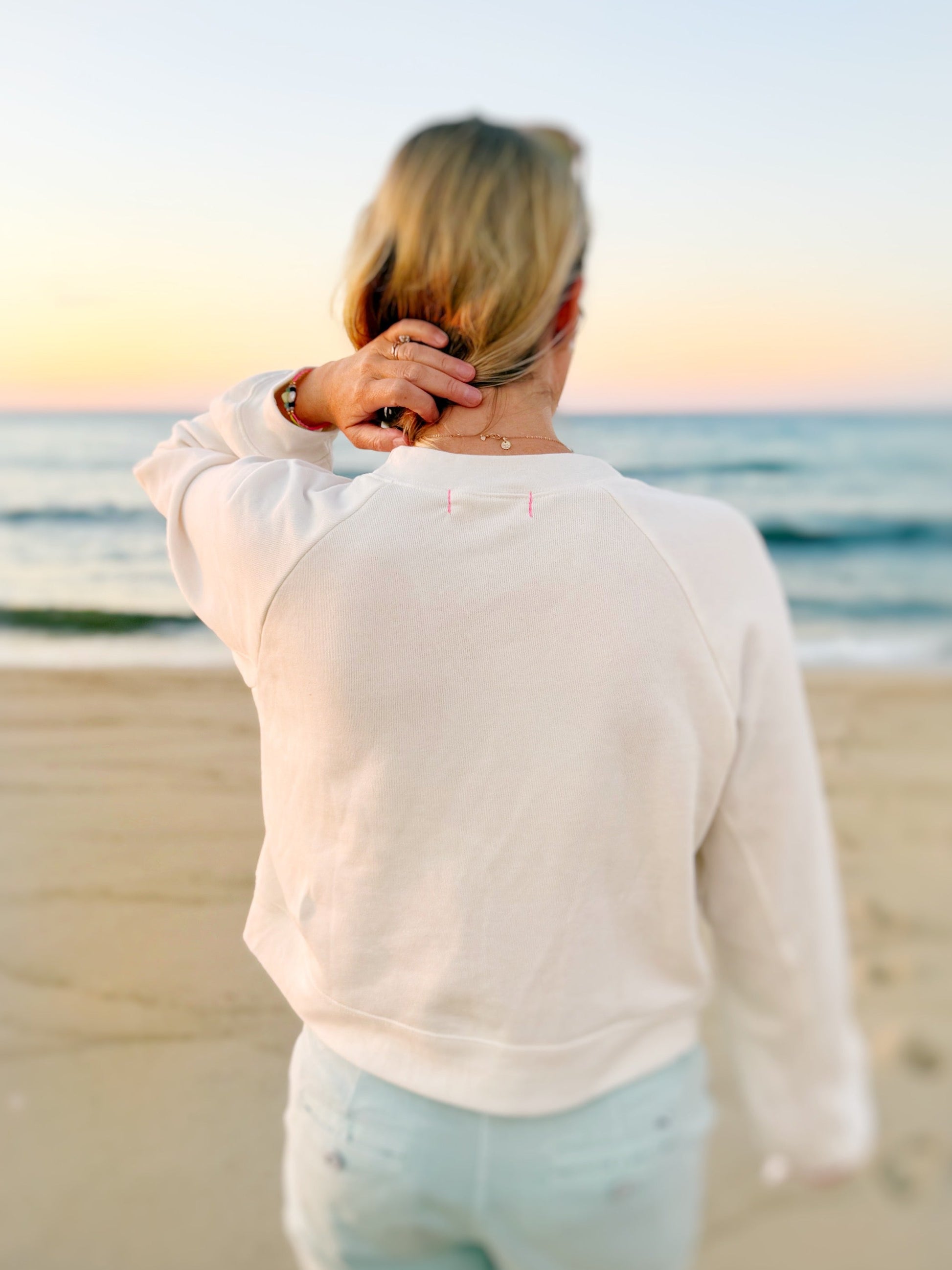 A woman viewed from behind on a beach at sunset, wearing a cream-colored long-sleeve sweatshirt and light blue shorts, with her hand near her neck.