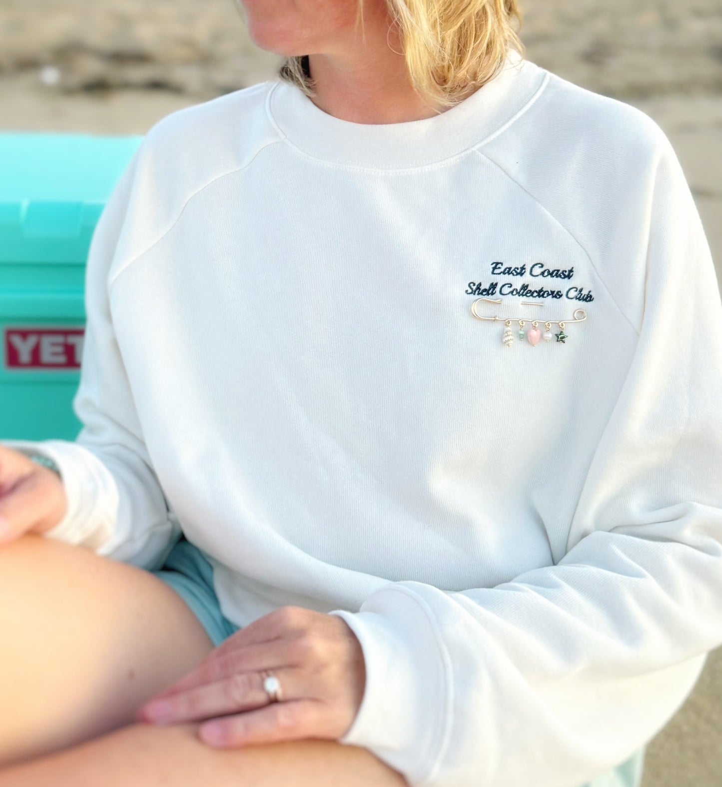 A person wearing a white crewneck sweatshirt with 'East Coast Shell Collectors Club' embroidered on the chest, sitting outdoors near a turquoise cooler.