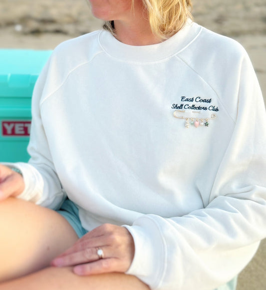 A person wearing a white crewneck sweatshirt with 'East Coast Shell Collectors Club' embroidered on the chest, sitting outdoors near a turquoise cooler.