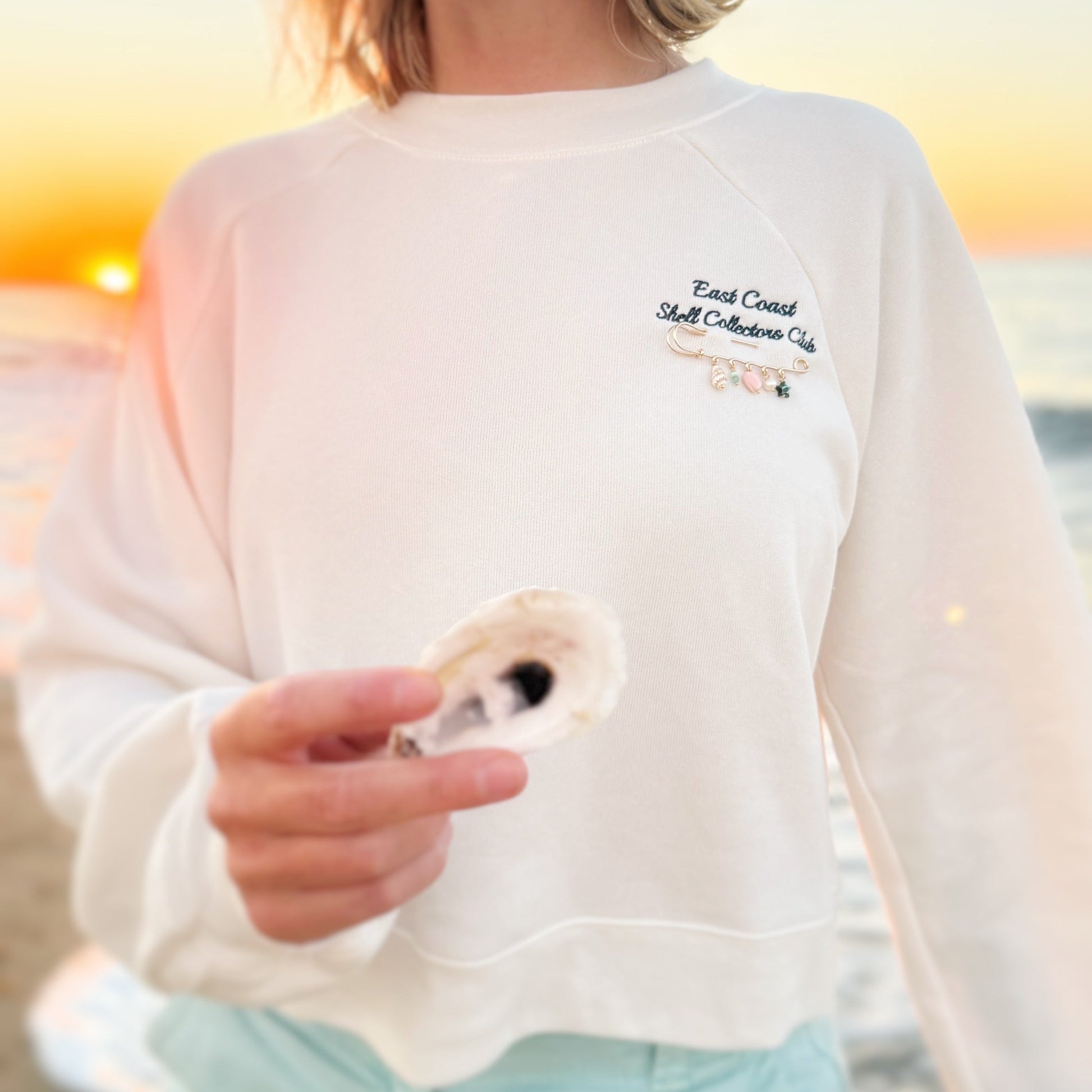 A person on a beach at sunset wearing a white crewneck sweatshirt with 'East Coast Shell Collection Co' embroidered on the chest, holding a seashell.