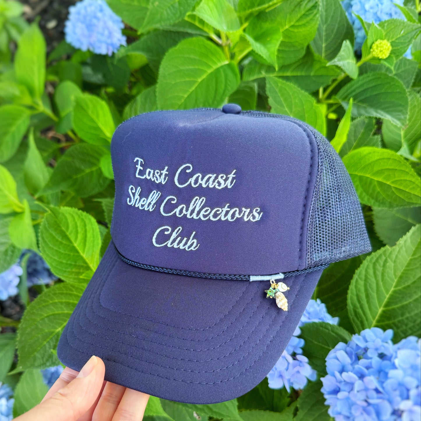 A navy blue trucker hat with 'East Coast Shell Collectors Club' embroidered in white on the front, held by a hand against a background of green hydrangea flowers.