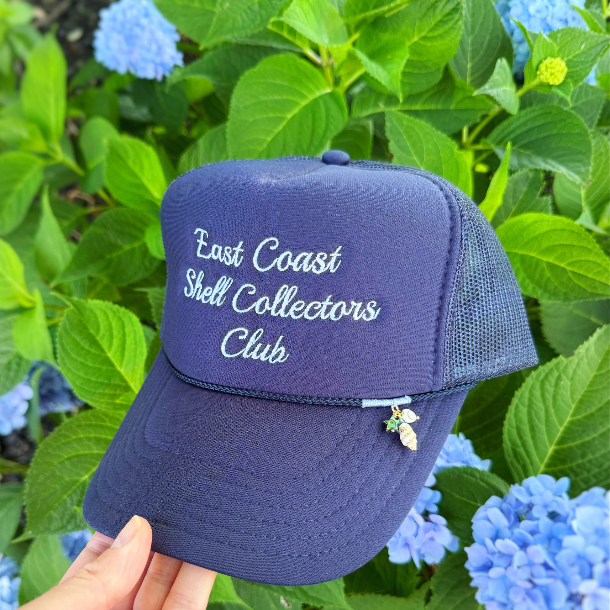 A navy blue trucker hat with 'East Coast Shell Collectors Club' embroidered in white on the front, held by a hand against a background of green hydrangea flowers.