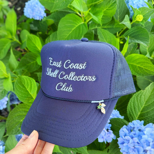A navy blue trucker hat with 'East Coast Shell Collectors Club' embroidered in white on the front, held by a hand against a background of green hydrangea flowers.