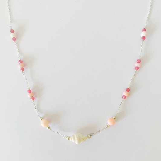 the pink lemonade shellebration necklace by mermaids and madeleines has a shell and coral beads at the center with stations of pink opal and pink tourmaline spread throughout the sterling chain to the clasp. this necklace is pictured on a white surface