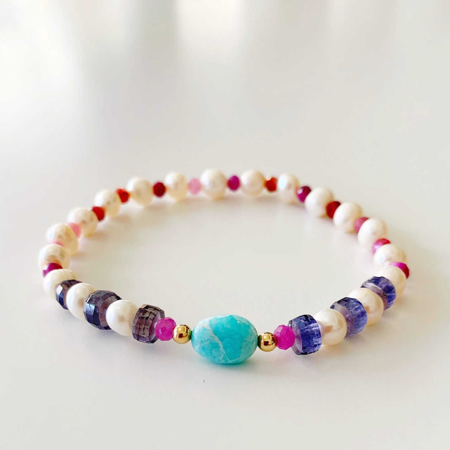 the sunset cruise stretch bracelet by mermaids and madeleines is a once of a kind created with amazonite in the center with 14k gold filled beads and then complimented by freshwater pearls, iolite tire beads and natural ruby faceted rondelles. this bracelet is photographed flat on a white surface