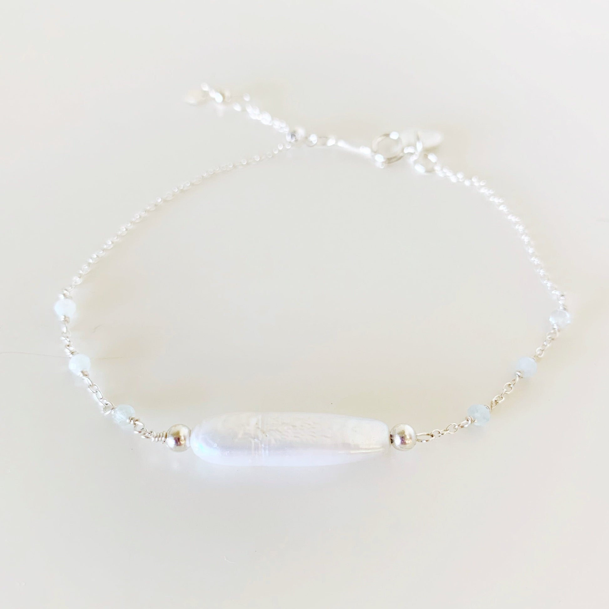 the truro bracelet by mermaids and madeleines is a sterling silver chain based adjustable bracelet with a freshwater stick pearl at the center. there are microfaceted aquamarine gems wired into the chain on either side and there's a slide bead near the clasp to tighten the bracelet. this piece is photographed from the front on a white surface