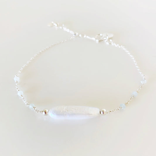 the truro bracelet by mermaids and madeleines is a sterling silver chain based adjustable bracelet with a freshwater stick pearl at the center. there are microfaceted aquamarine gems wired into the chain on either side and there's a slide bead near the clasp to tighten the bracelet. this piece is photographed from the front on a white surface