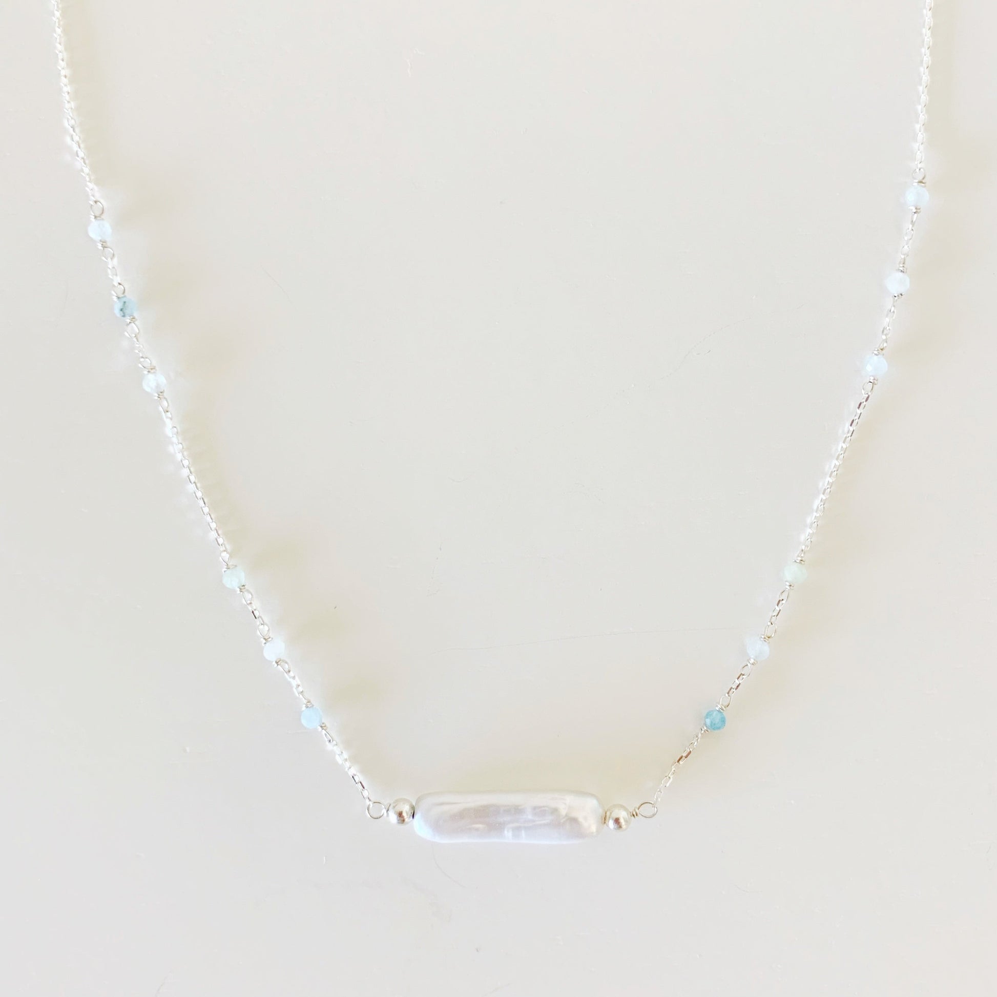 the truro necklace by mermaids and madeleines is created with sterling silver chain dotted with microfaceted aquamarine gems and has a freshwater stick pearl at the center. this is a closer up view of the necklace and photographed on a white surface