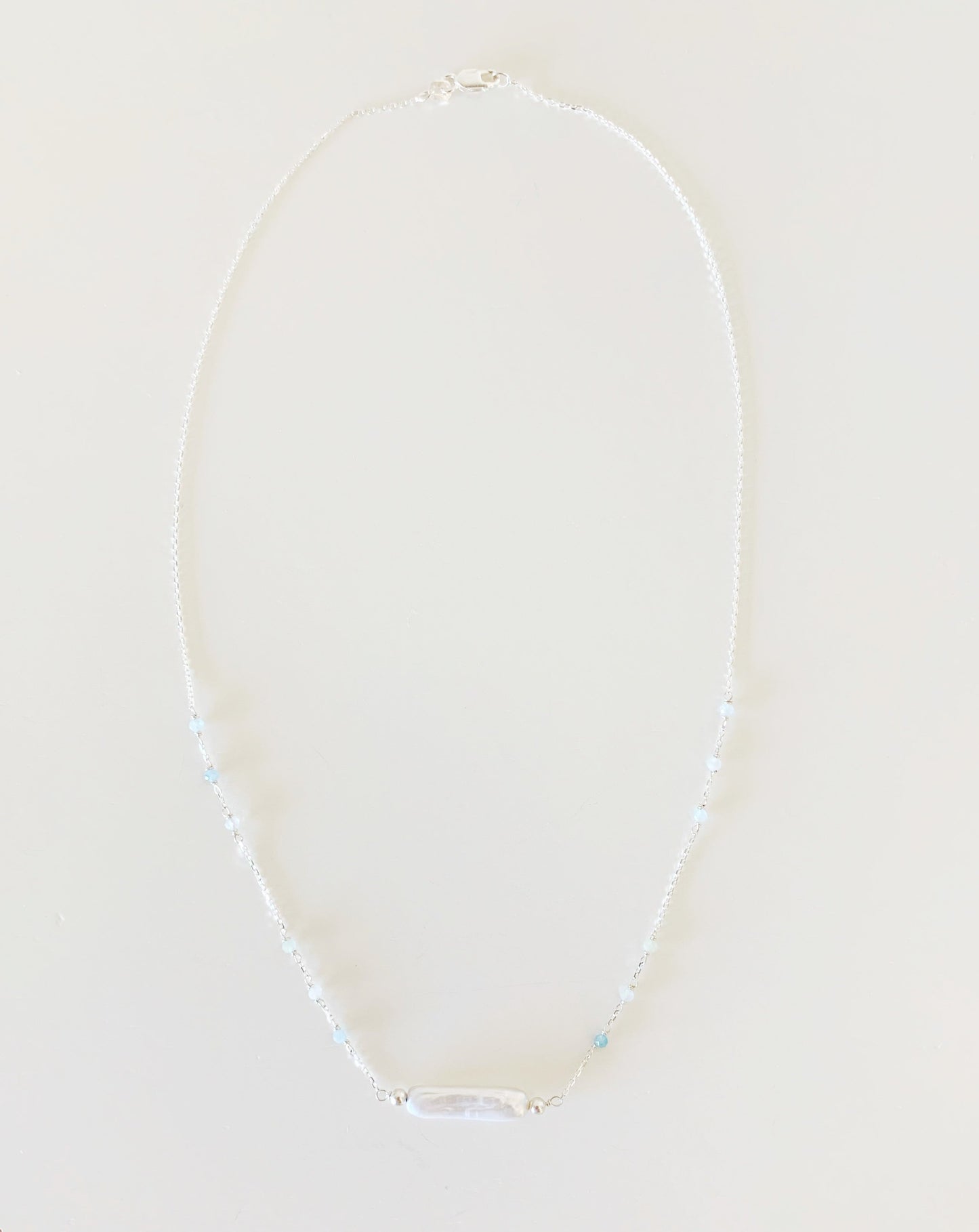the truro necklace by mermaids and madeleines is created with sterling silver chain dotted with microfaceted aquamarine and with a freshwater stick pearl at the center. this is a full view of the necklace photographed on a white surface