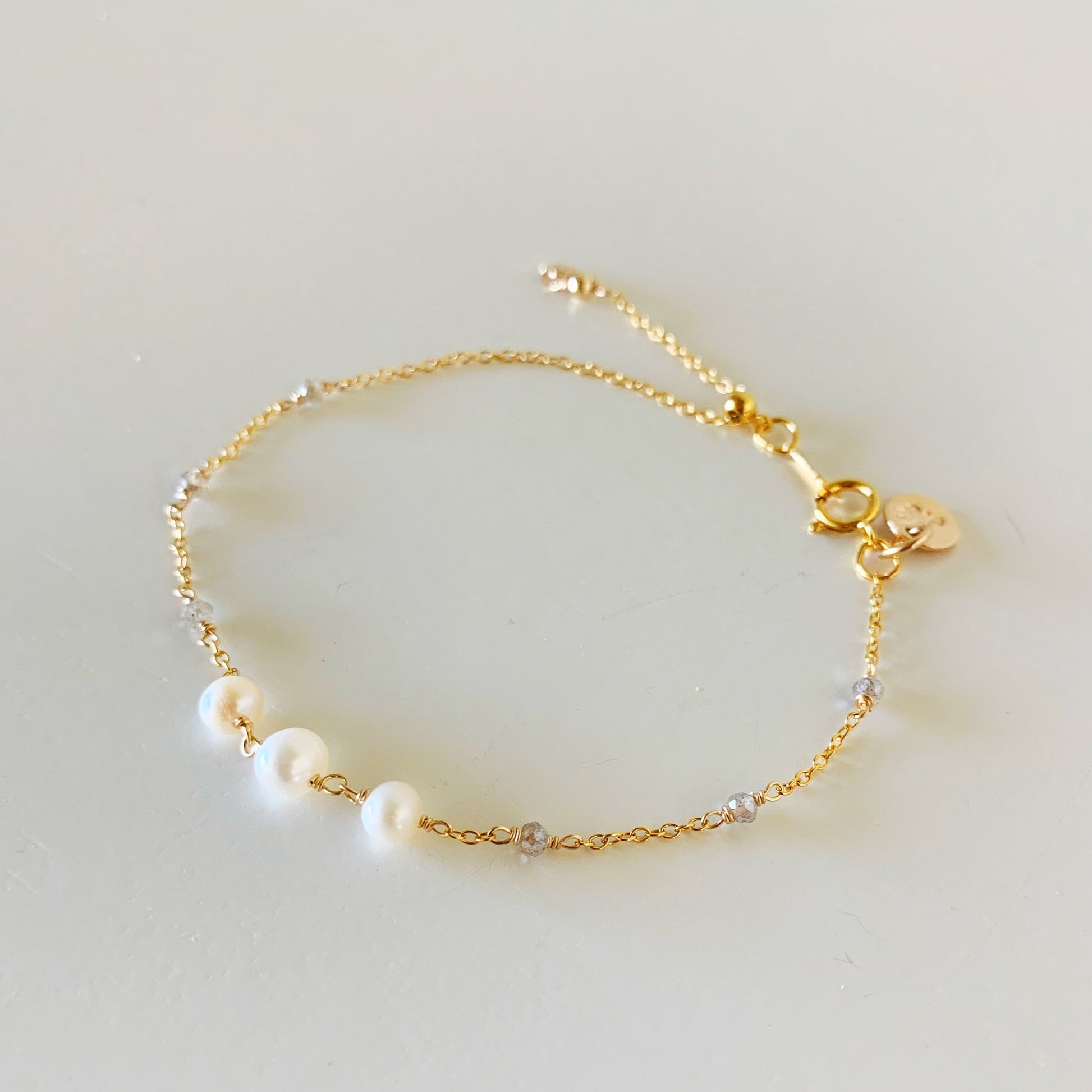 the westport bracelet by mermaids and madeleines is a 14k gold filled chain based design with an adjustable slide bead near the clasp to tighten the bracelet. this piece features 3 freshwater pearls at the center and has tiny microfacet labradorite beads on the sides of the chain. this bracelet is facing slightly left and photographed from a top down angle on a white surface