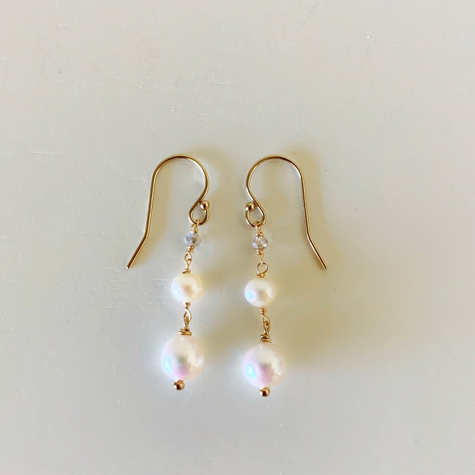 the westport earrings by mermaids and madeleines are a simple dangling style with freshwater pearls graduating to a small microfaceted labradorite bead all hanging from 14k gold filled earring findings. this pair of earrings is photographed flat on a white surface