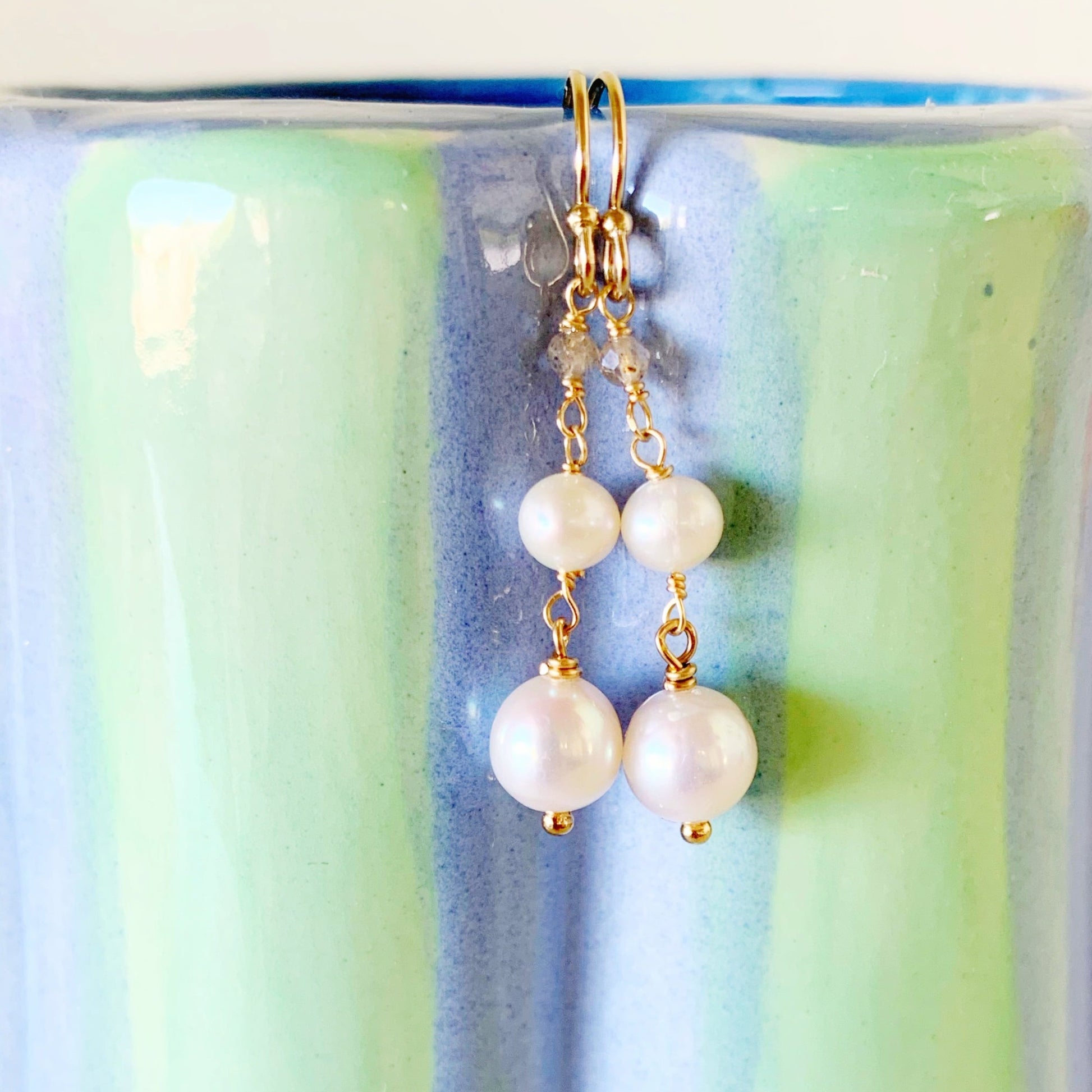 the westport earrings by mermaids and madeleines are handwrapped freshwater pearl ,tapered linear earrings available in sterling silver or 14k gold filled, and complimented by tiny microfaceted labradorite rondelles. this pair photographed has 14k gold filled wire and findings and is resting on a light blue and green jar.