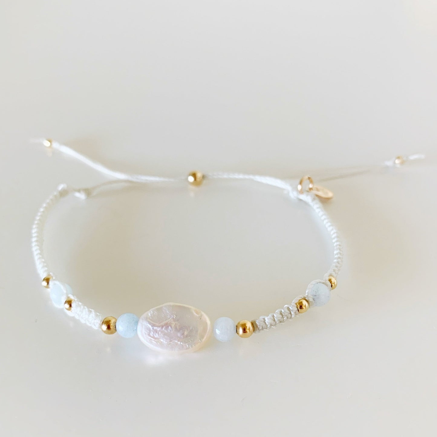 the world is your oyster bracelet by mermaids and madeleines is designed with white shell color cord with freshwater oval pearl at the center with aquamarine beads on either side. this bracelet is photographed from the front on a white surface