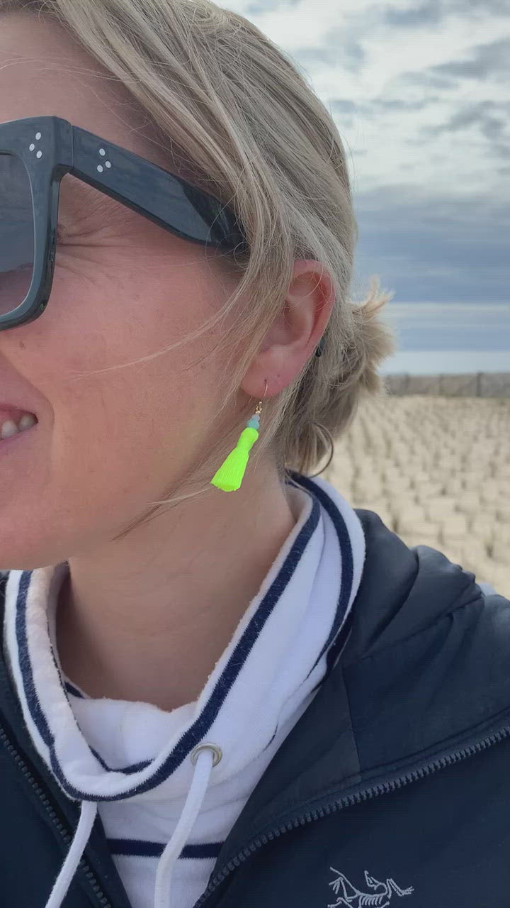 Captiva tassel earrings are created with neon yellow cord and topped with amazonite and blue lace agate beads with 14k gold filled findings. This is a close up video of the earring on a woman and the tassels are blowing in the wind at the beach