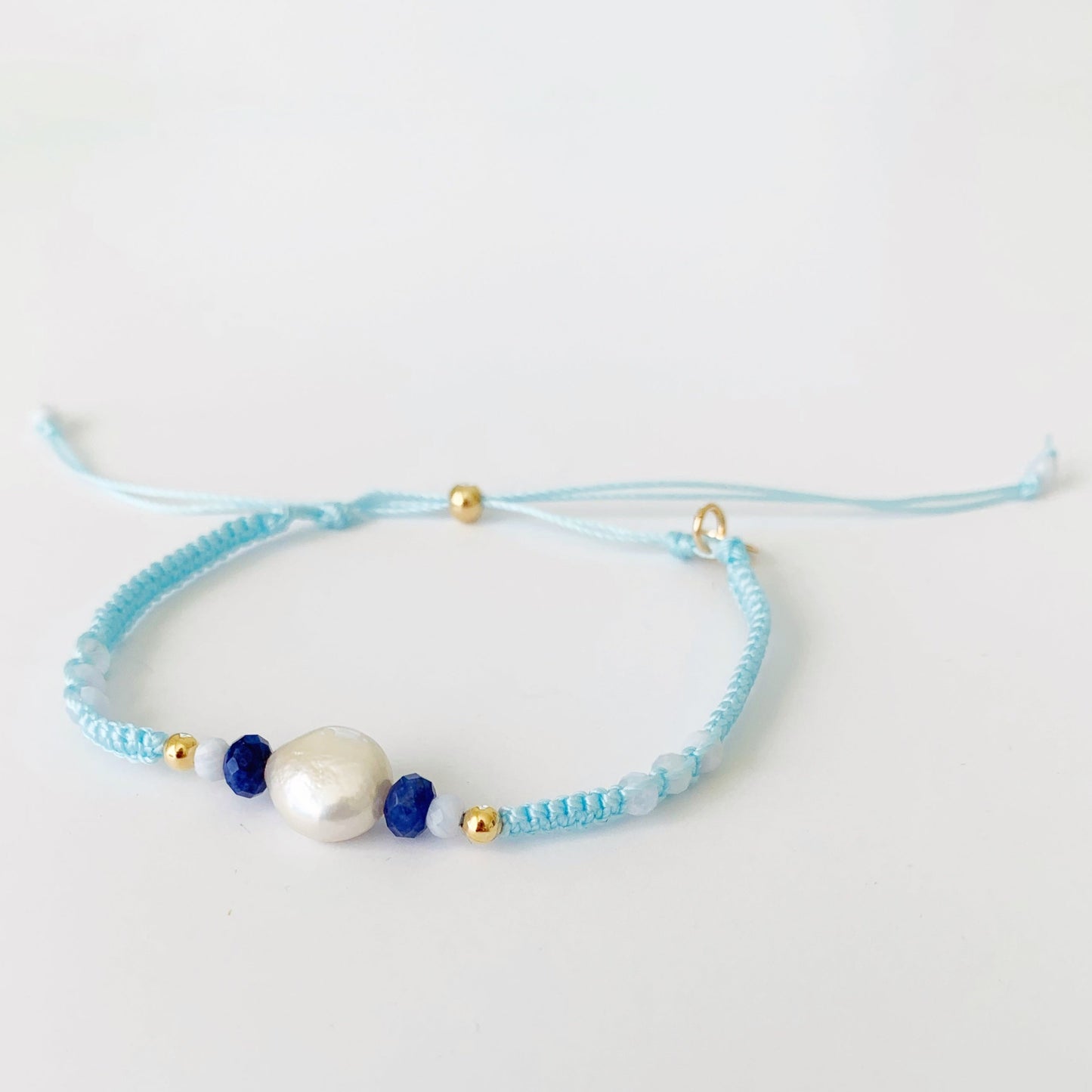 The forget me knot macrame bracelet is a light blue friendship bracelet with a freshwater coin pearl, and semiprecious beads at the center with a 14k gold filled slide bead clasp. this bracelet is pictured on a white surface