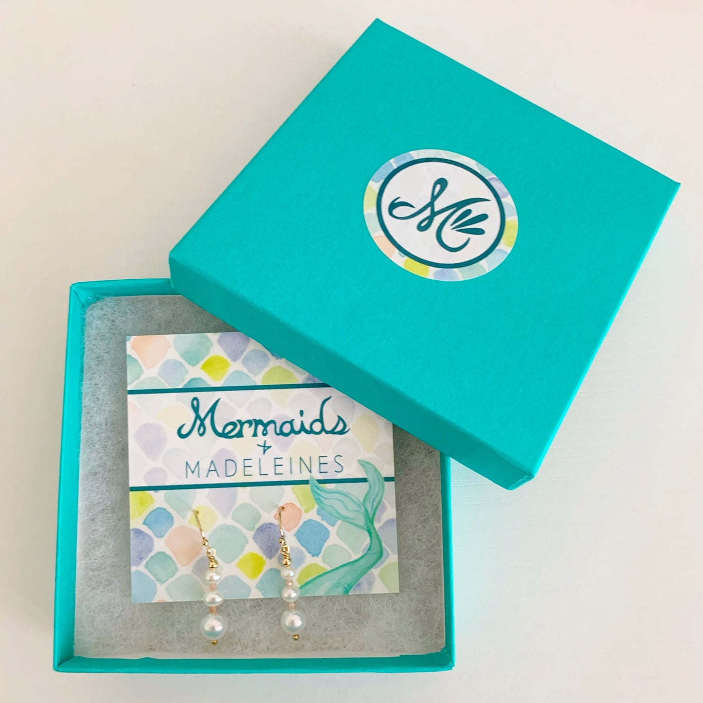 barrington earrings by mermaids and madeleines feature freshwater pearls in a linear taper, with peach moonstone accents and 14k gold filled findings. This pair of earrings is carded and placed in a cotton lined teal gift box on a white surface