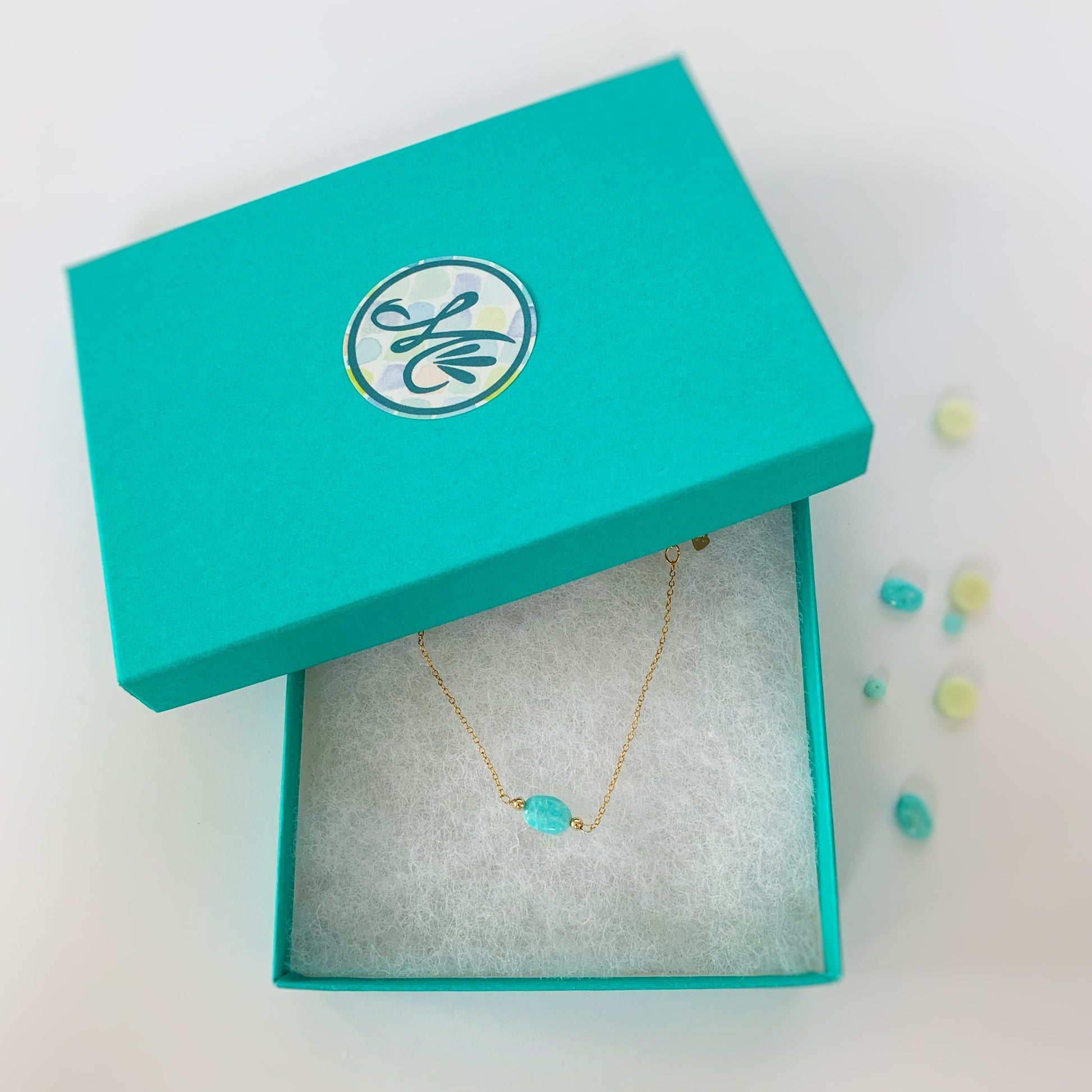 Boxed Laguna adjustable bracelet. This laguna bracelet is created with 14k gold filled chain and findings and a bright aqua oval shaped amazonite bead at the center of the bracelet. This bracelet is pictured in a cotton lined teal gift box with an M mermaids and madeleines logo sticker on the lid. The item is photographed on a white background