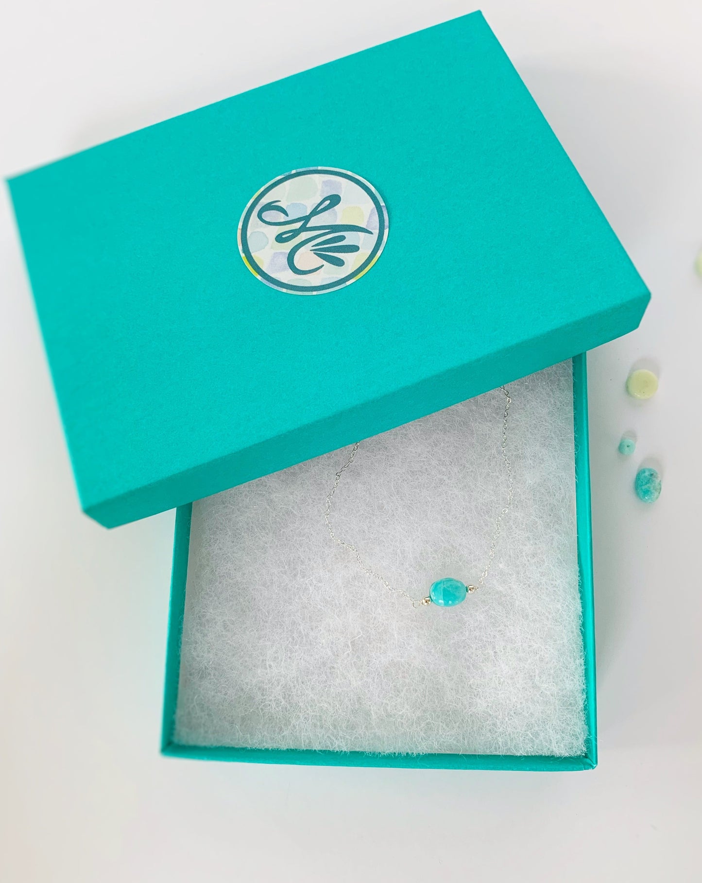 Boxed picture of the sterling silver adjustable laguna bracelet. The laguna bracelet has sterling silver findings and chain with a bright aqua, oval shaped amazonite bead at the center of the bracelet. This one is pictured in a cotton lined teal gift box with an M mermaids and madeleines logo on the lid. The item is photographed on a white background