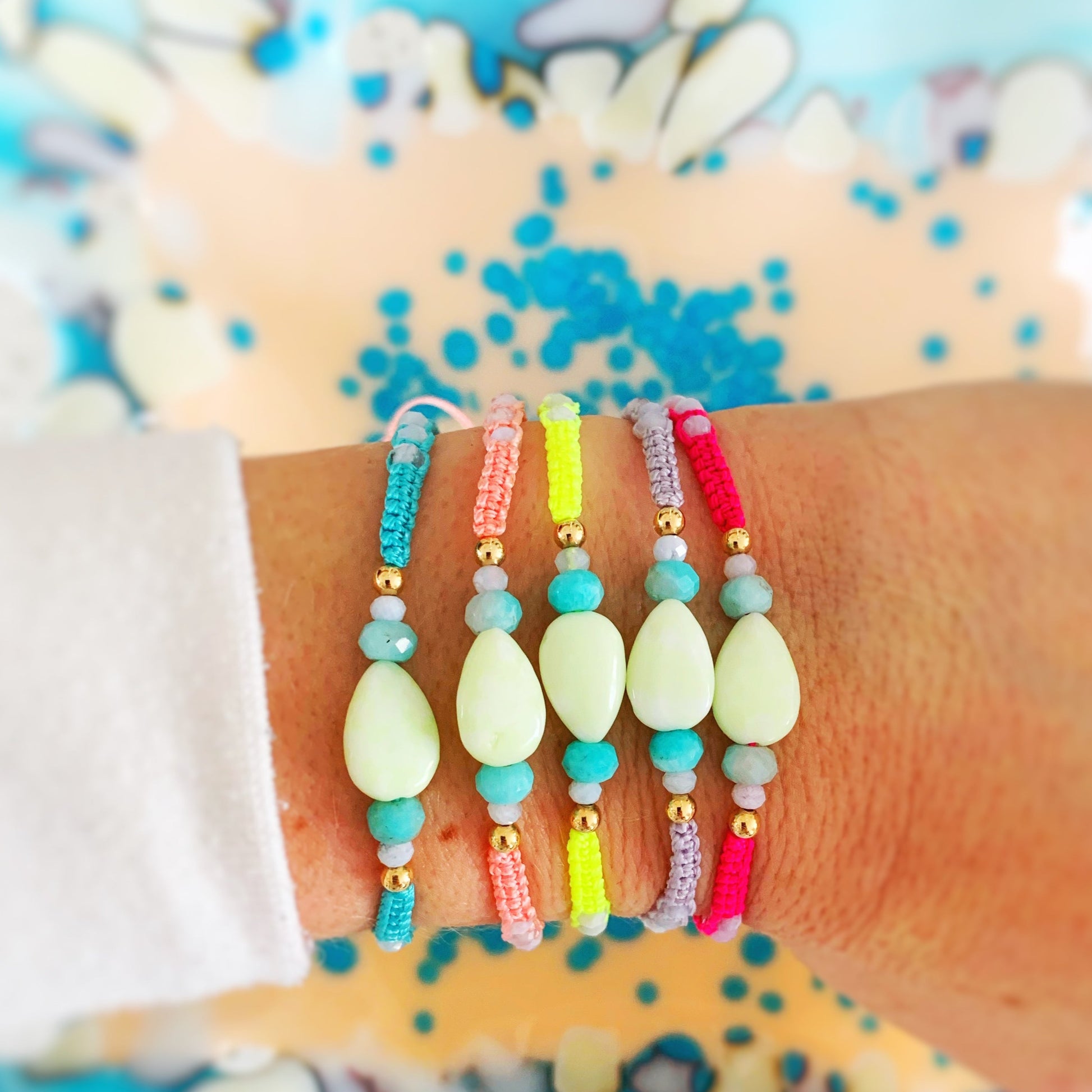 All 5 color options of the Captiva macramé bracelets worn on a wrist, in colors blue, peach, yellow, lavender, pink bracelets worn on a wrist stacked and photoed over a blue and white glass dish 