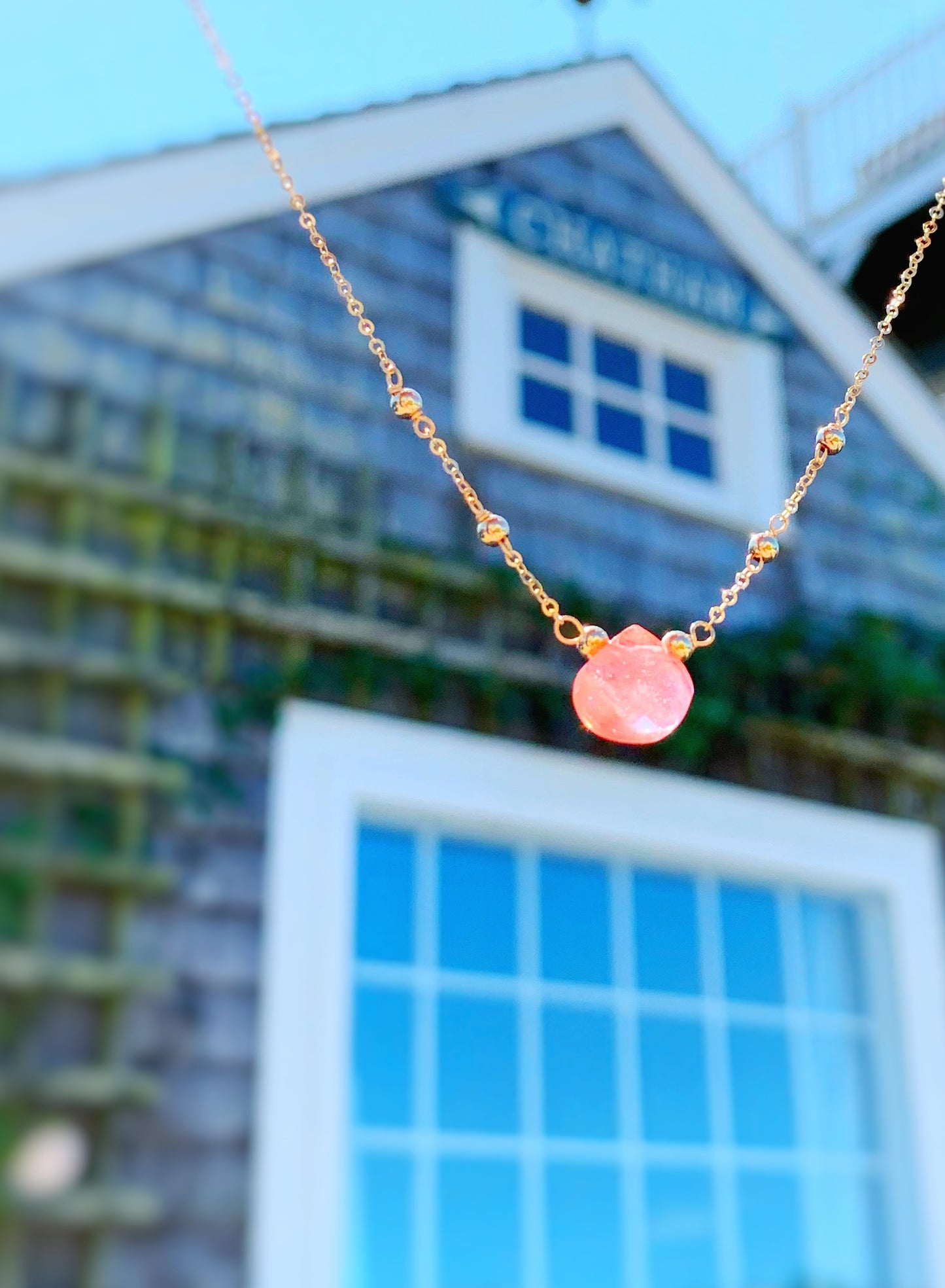 Chatham Necklace by Mermaids and Madeleines features a cherry quartz briolette drop and 14k gold filled findings, beads and chain. This one is pictured in the air with a blurred shingled beach cottage in the background