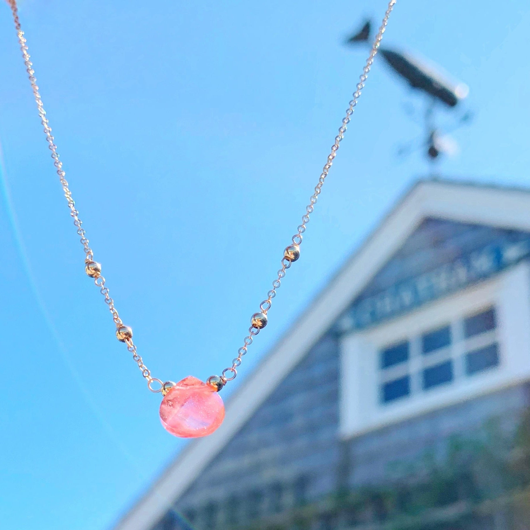 Chatham Necklace by Mermaids and Madeleines features a cherry quartz briolette and 14k gold filled beads, findings and chain. This one is photographed in the air with a blurred shingled beach cottage in the background