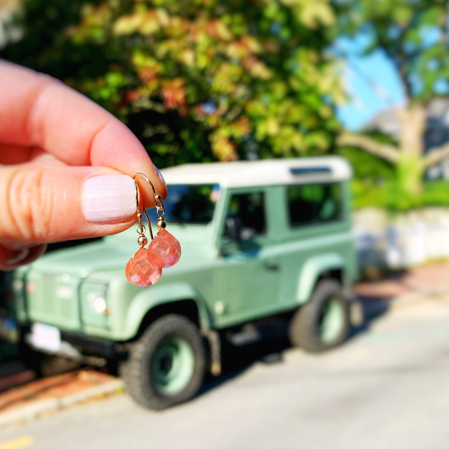 Chatham Earrings by Mermaids and Madeleines feature cherry quartz drops and 14k gold filled findings. this pair is held up in a hand in front of a green offroading vehicle as a background