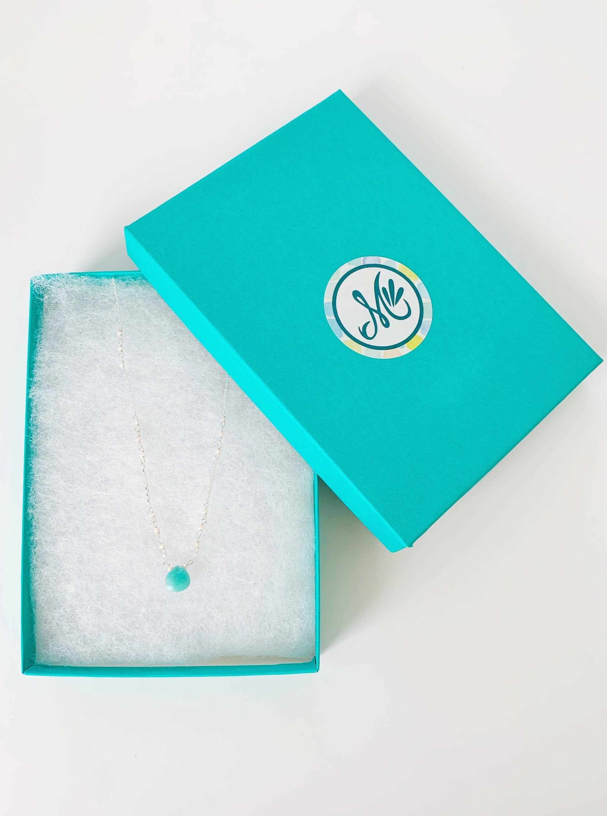 The island air necklace by mermaids and madeleines is pictured here in a cotton lined teal gift box. The necklace has an amazonite gem at the center and freshwater pearl in the sterling silver chain