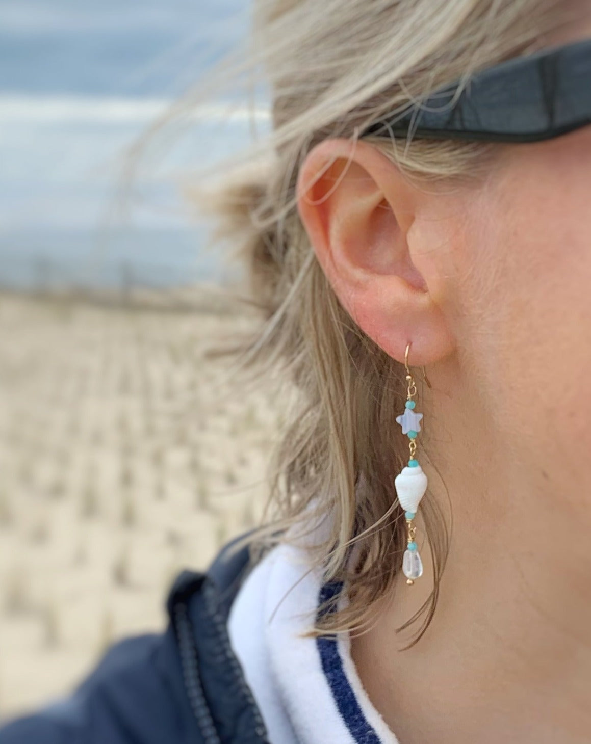 The Island Hopper earrings are photographed close up on a woman at the beach with focus on the earring The earrings are designed to be slightly asymmetrical. The earrings are created with 14k gold filled findings and wire, both end with an irregular shape of rose quartz, have a shell in the center of the earring but the first bead on each earring is different. One has a star shaped blue lace agate bead, and the other started with a rough faceted Peruvian opal rondelle. 