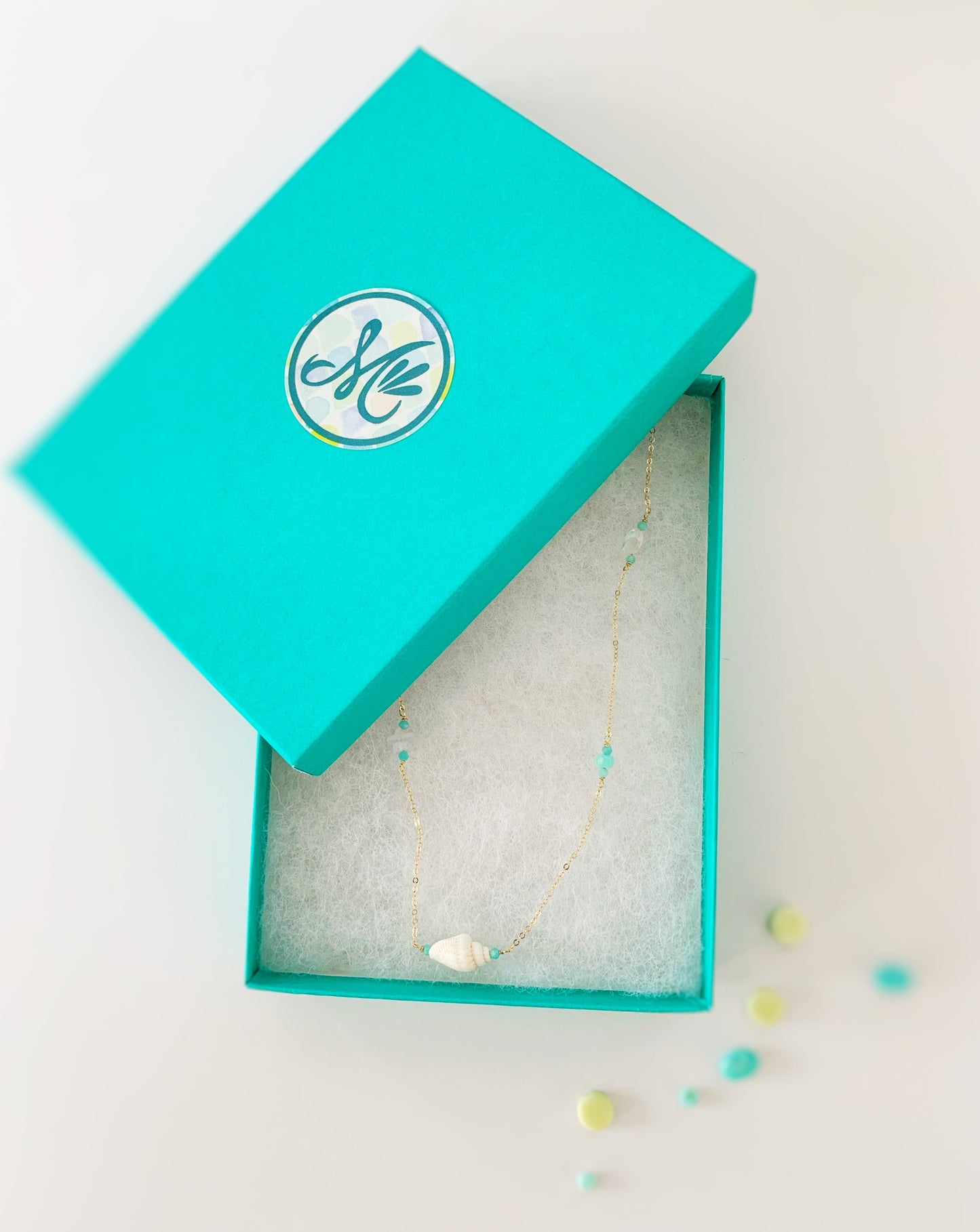 The Island hopper is from the shellebration collection, its pictured here in a cotton lined teal color gift box with a mermaids and madeleines logo sticker on it. the necklace is in the box and it's photographed on a white background