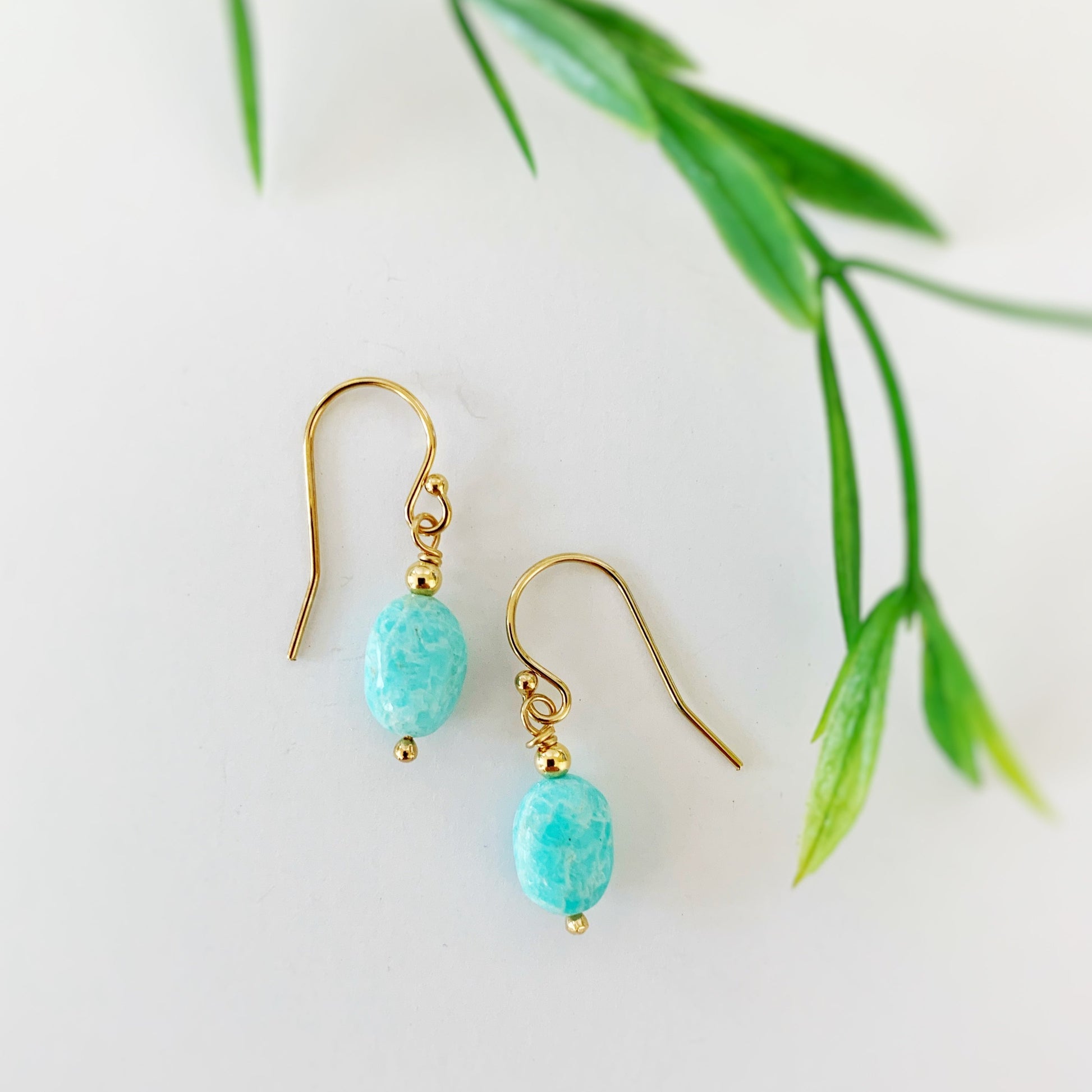 A picture of the laguna earrings in 14k gold filled. The laguna earrings are created with 14k gold filled beads and findings and bright aqua oval shaped amazonite beads. This pair is photographed on a white background with blurred greenery on the right side of the picture