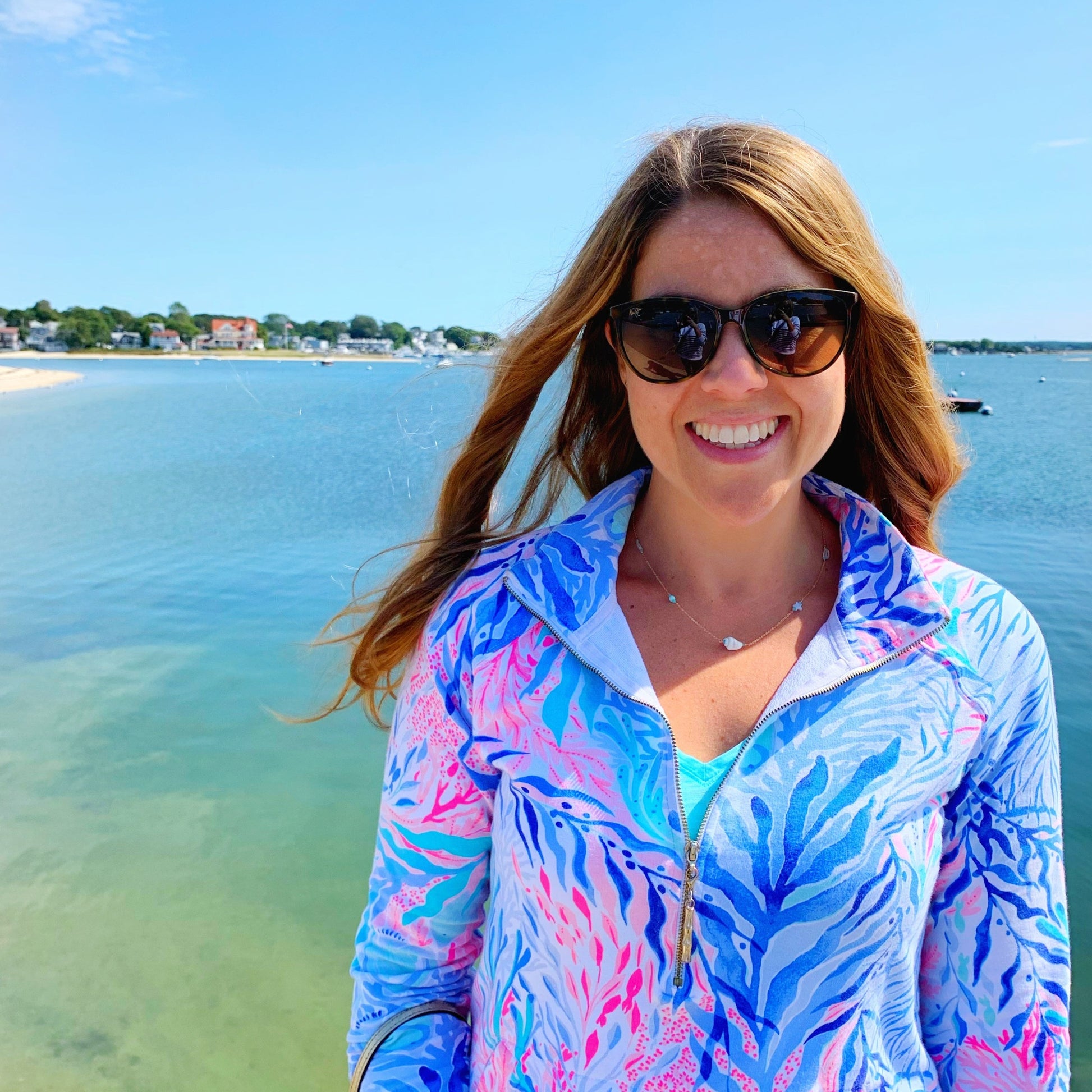 Model dressed in bright lilly pulitzer 3/4 zip up top is wearing the mermaids and madeleines island hopper necklace that is created with semiprecious gems and a shell at the cetner. the model has onset beach behind her