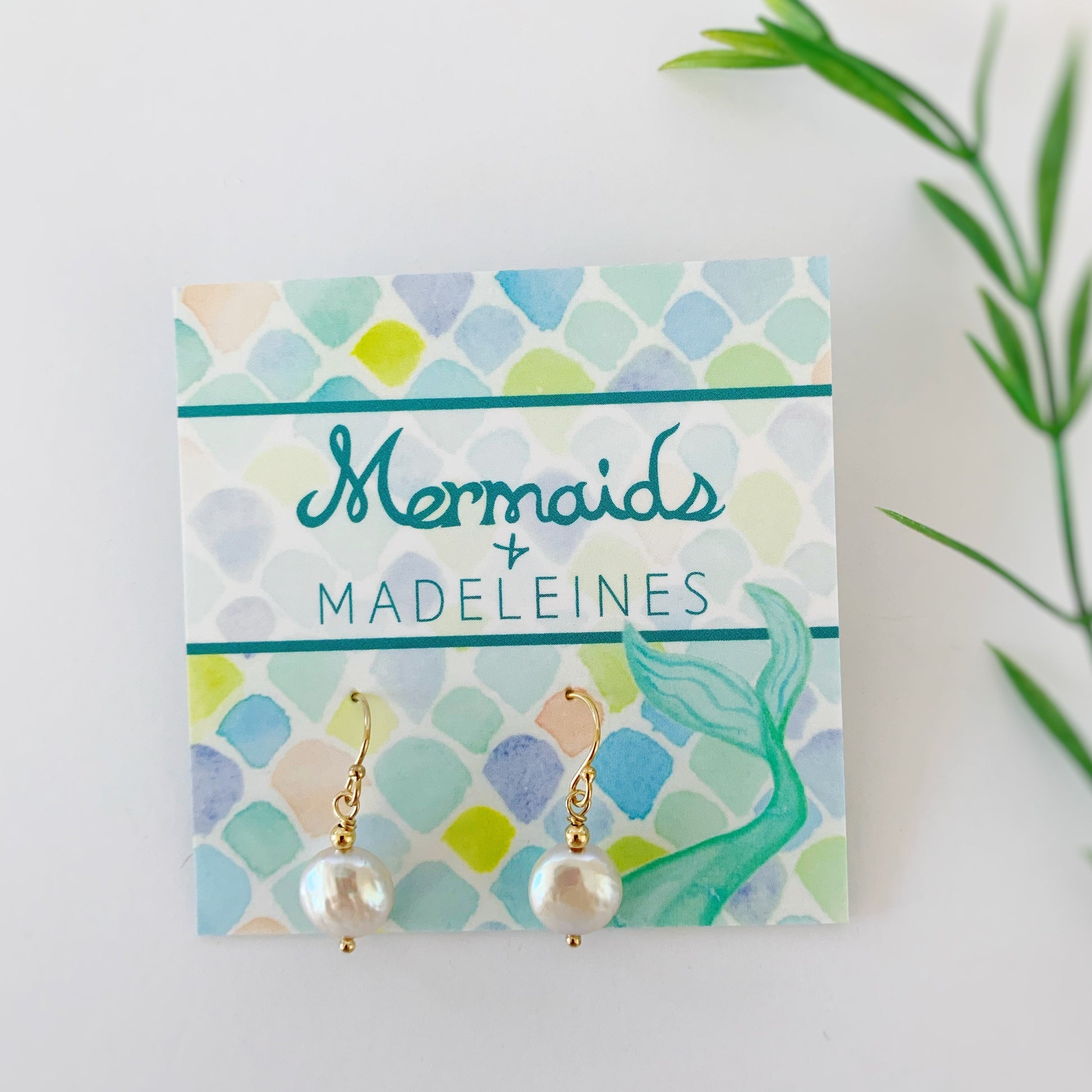 Newport Earrings in 14k gold filled. This pair of earrings is created with 14k gold filled beads and findings and iridescent freshwater coin pearls on a mermaids and madeleines earring card photographed on a white background with blurred greenery on the right side for interest