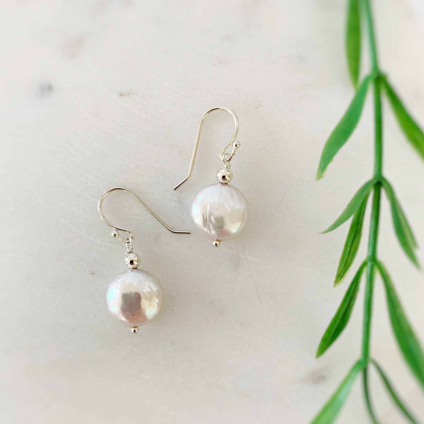 Newport earrings in sterling. This pair of earrings is created from sterling silver beads and earring findings with iridescent freshwater coin pearls. This pair is photographed on a white background with blurred greenery along the right side of the photo