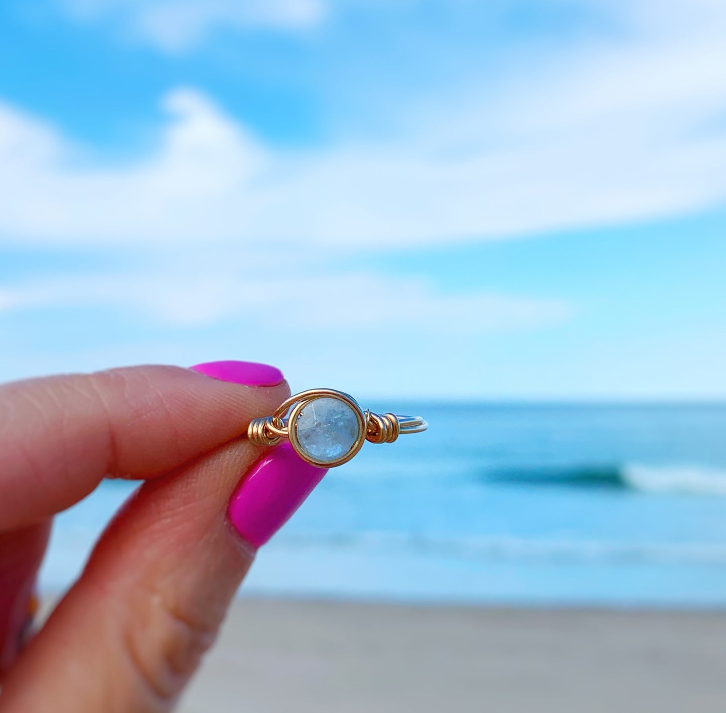 The raindrop ring featuring a coin shaped aquamarine bead is carefully wrapped in 14k gold filled wire into a wearable ring. this ring is being held up between fingers in front of a beach background
