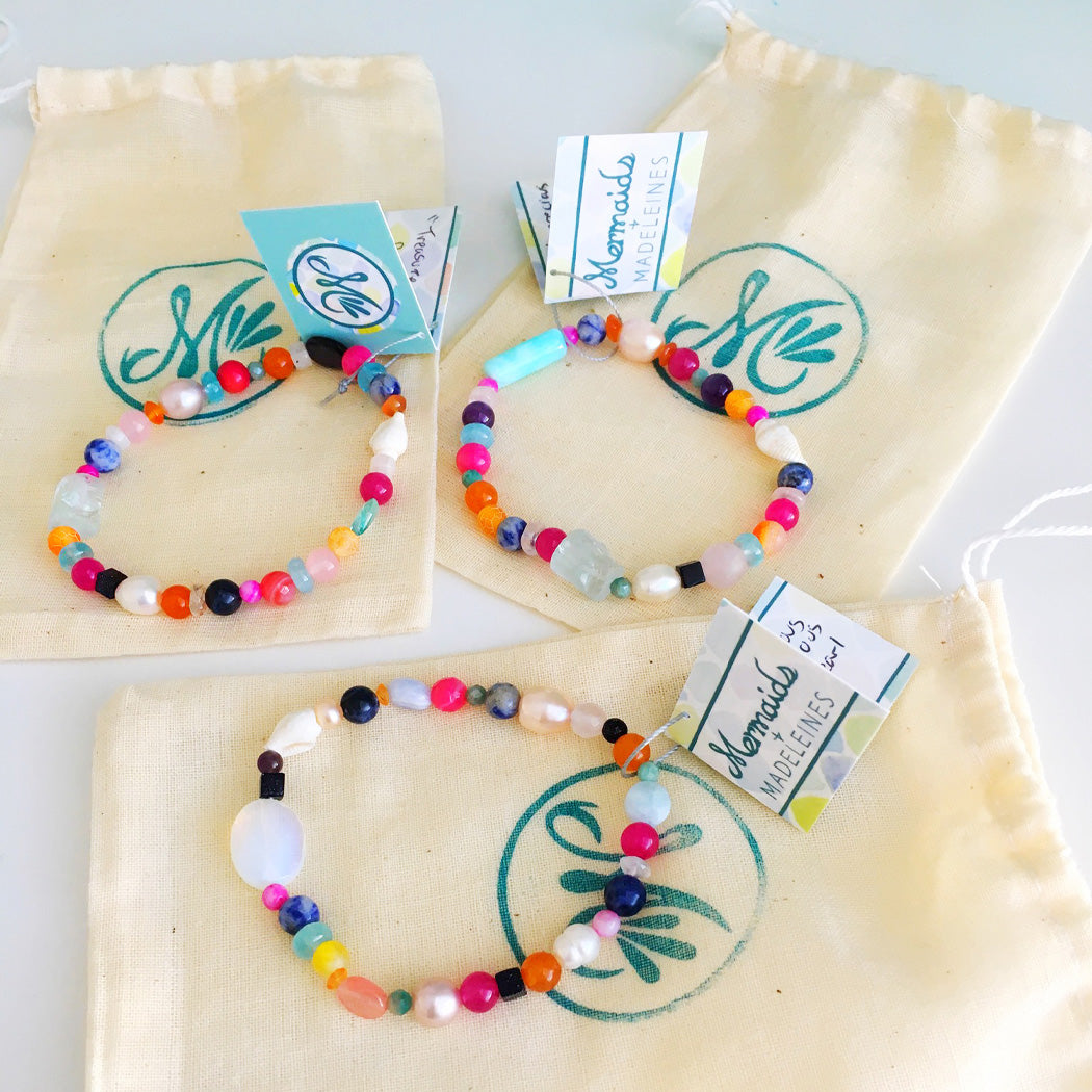 Treasure bracelets are stretchy bracelets made for adult women. The bracelets are created with bright precious and semiprecious stones, and freshwater pearls. Here 3 are pictured on top of the branded mermaids and madeleines pouch and on a white surface.