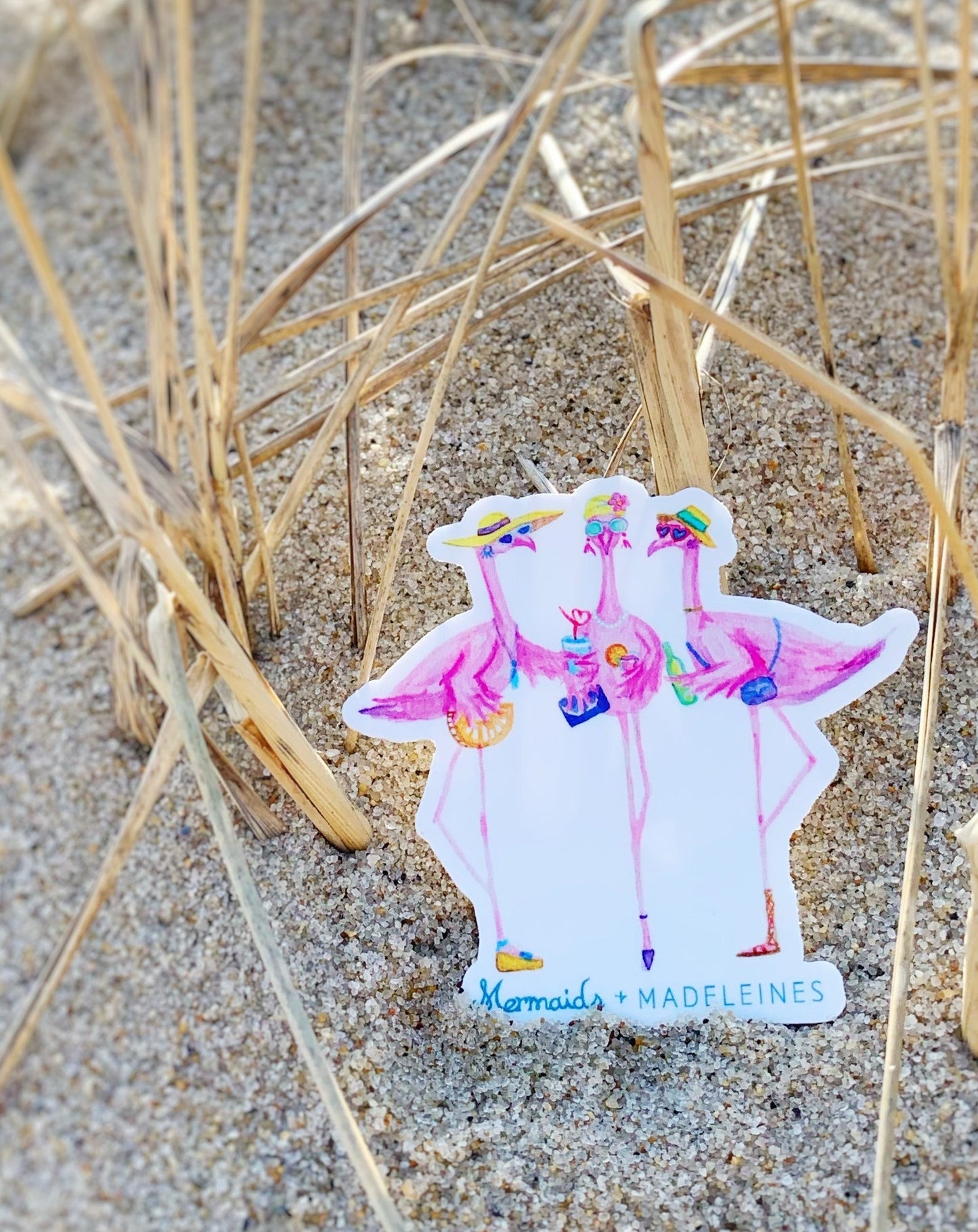 The flamingal sticker is created from original artwork made with acrylic paint and marker its 3 flamingo ladies dressed in summer hats and various footwear. This sticker is pictured sitting in the sand at the beach leaning against some beach grass
