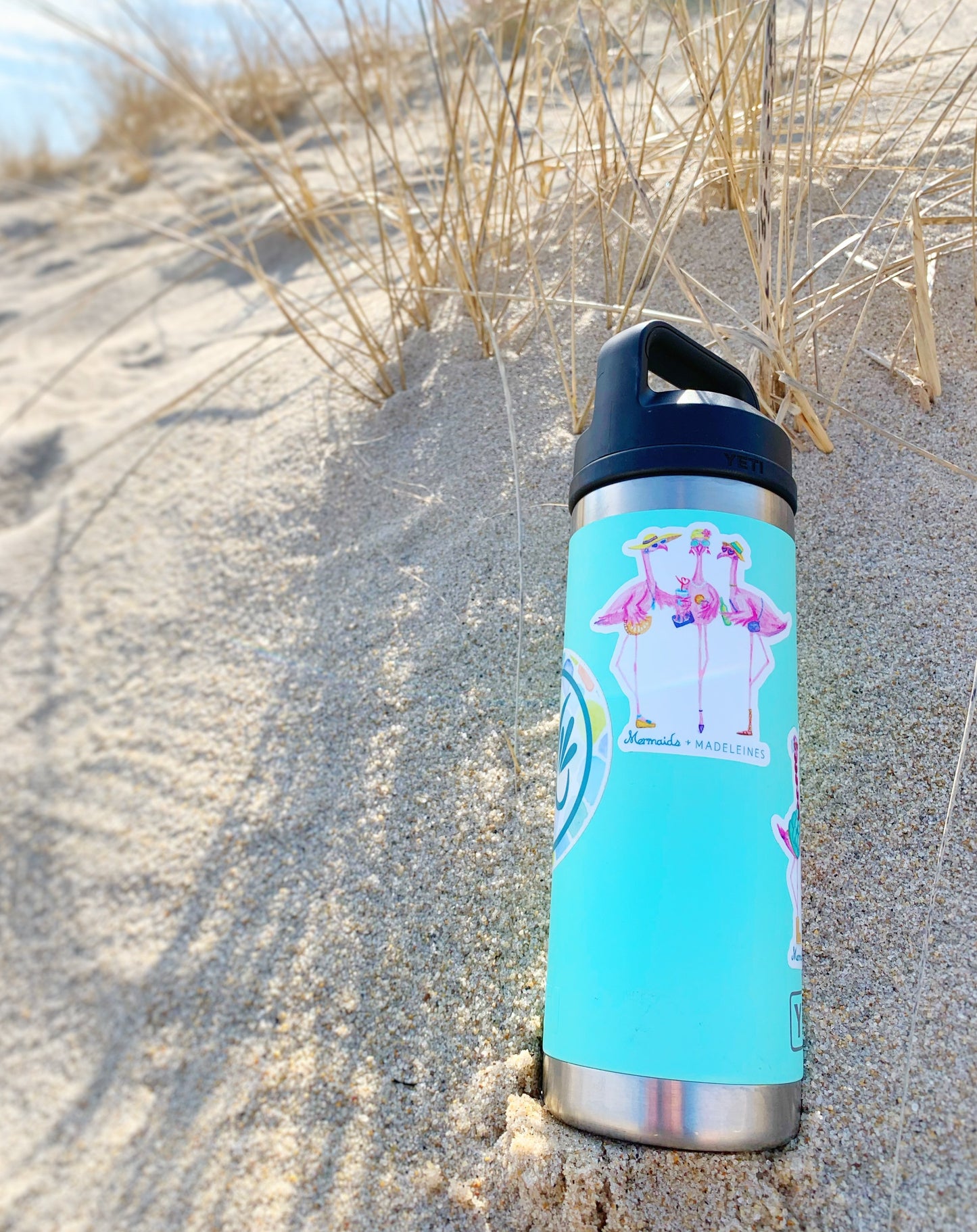 The flamingal sticker is created from original artwork made with acrylic paint and marker its 3 flamingo ladies dressed in summer hats and various footwear. This is a picture of a sticker on an aqua color yeti thermos sitting in the sand at the beach