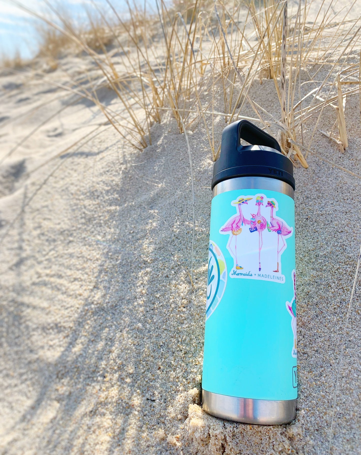 The flamingal sticker is created from original artwork made with acrylic paint and marker its 3 flamingo ladies dressed in summer hats and various footwear. This sticker is on a bright aqua Yeti thermos thats sitting in sand on the beach