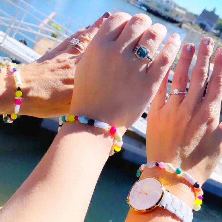 cropped photo of 3 friends with their arms extended, each wearing a treasure bracelet, a semiprecious, rainbow color stretchy bracelet with a shell. Just arms and hands are in view with the bracelets with a coastal background