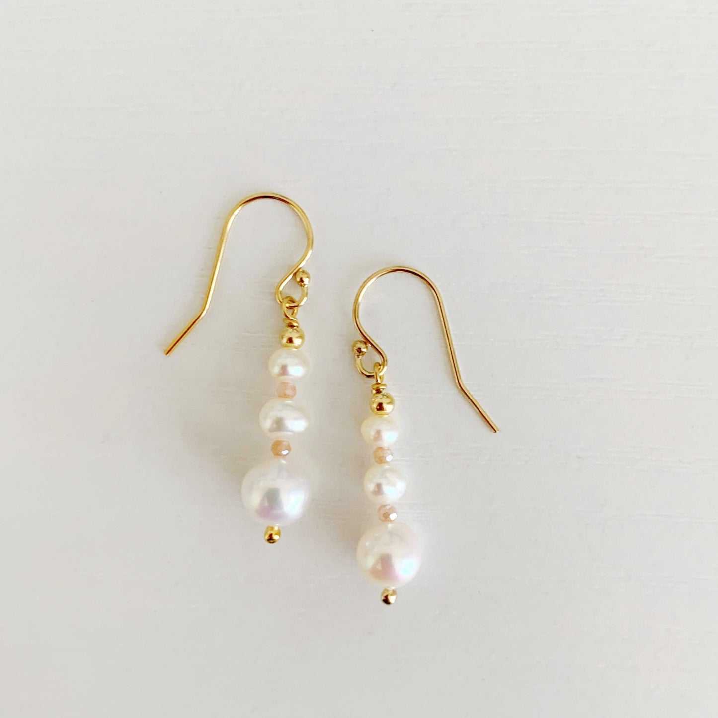 Barrington earrings by mermaids and madeleines feature freshwater pearls in a linear taper, with peach moonstone accents and 14k gold filled findings. this pair is photographed flat on a white surface with one earring placed slightly higher than the other on the surface