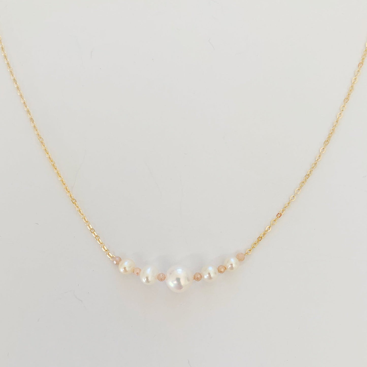 The barrington necklace by mermaids and madeleines has graduated freshwater pearls at the center with peach moonstone on 14k gold filled chain. this necklace is photographed close up and on a white surface
