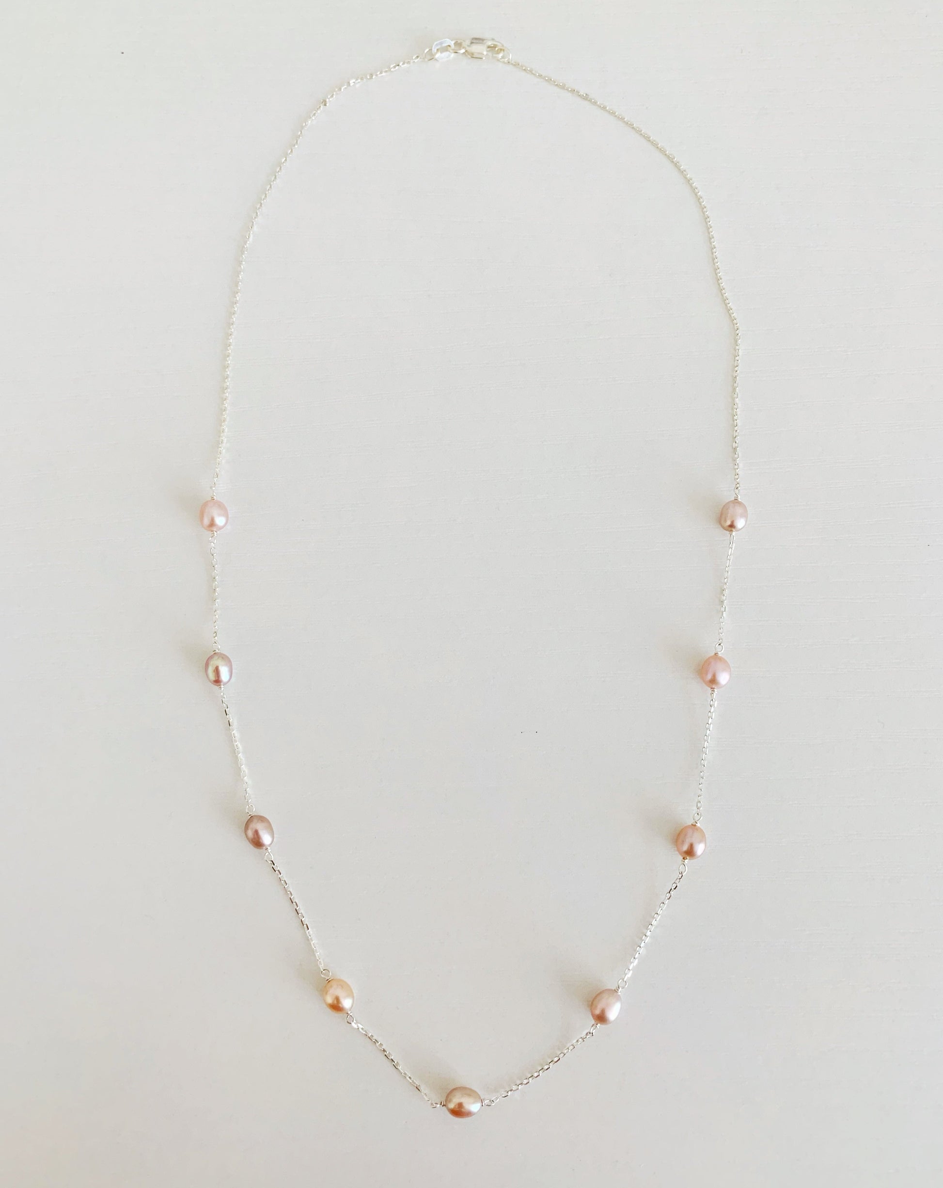 Block Island Necklace by mermaids and madeleines features natural color pink freshwater small oval pearls in stations on a sterling silver chain. there are 9 pearls spaced on the front half of the necklace. this is a photograph of the entire necklace on a white surface