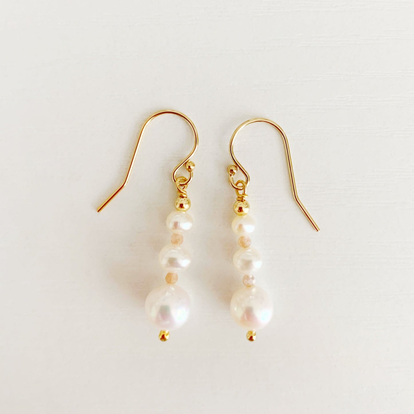 barrington earrings by mermaids and madeleines feature freshwater pearls in a linear taper with peach moonstone accents and 14k gold filled findings. this pair is photographed flat on a white surface
